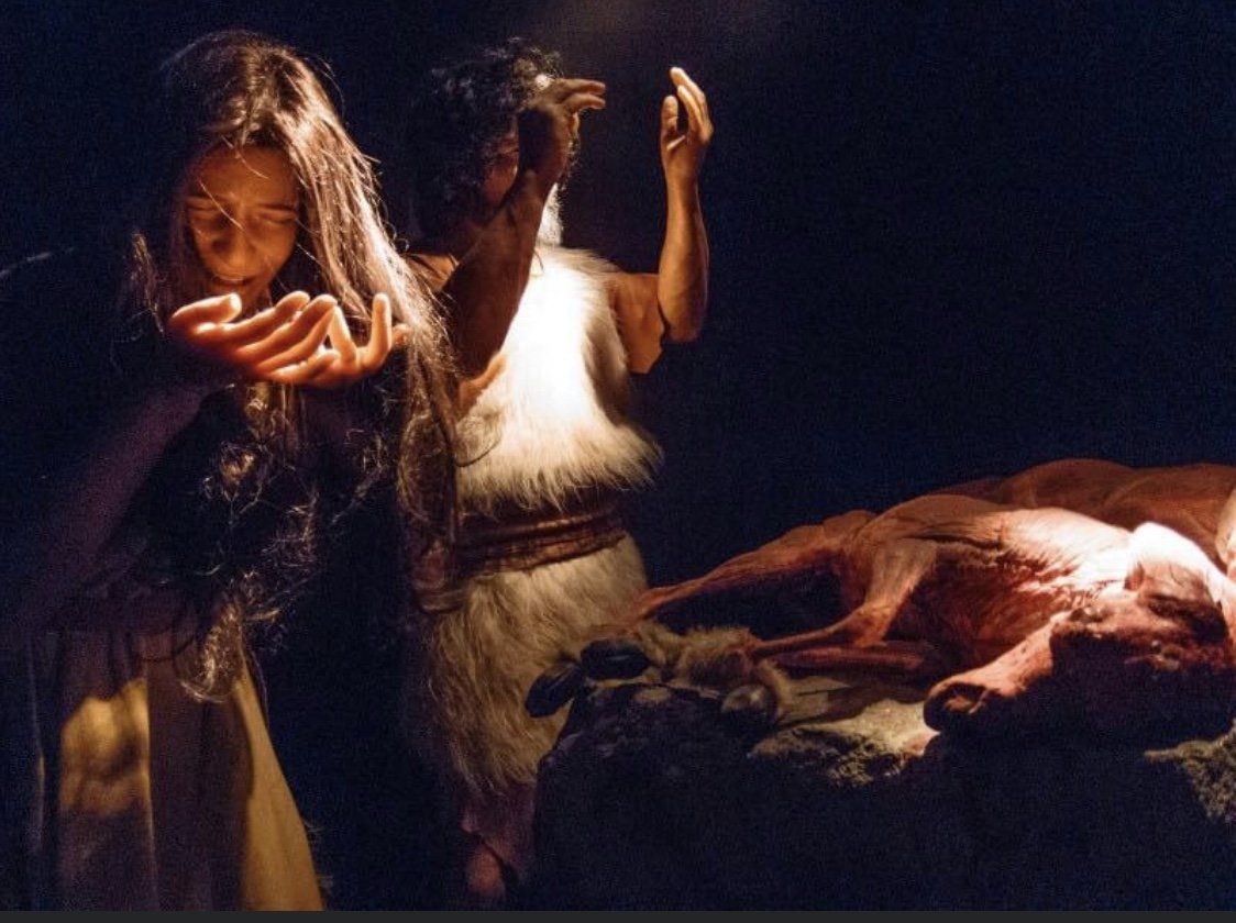 People in dim space, one holding an object to their face, another with hands raised, near a carcass on a stone.