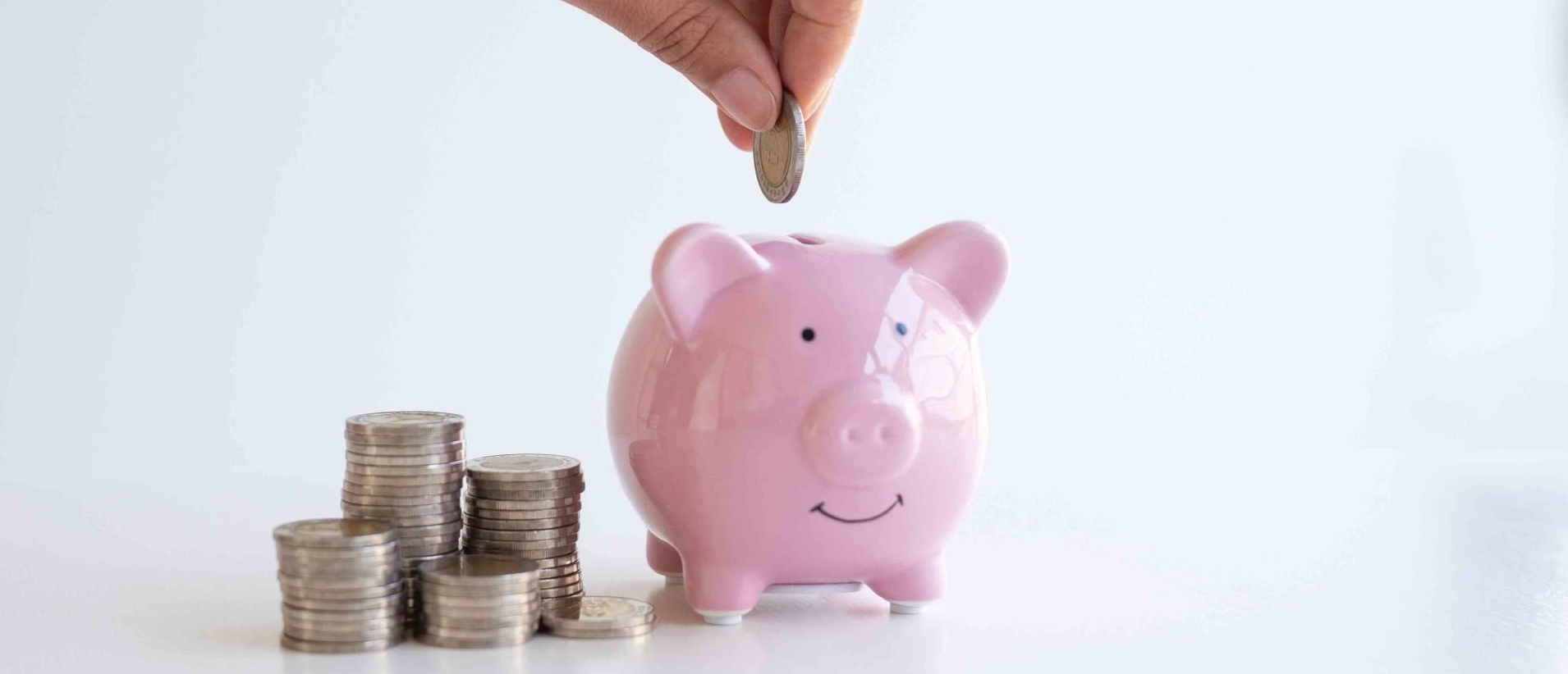 A person is putting a coin into a piggy bank next to stacks of coins.