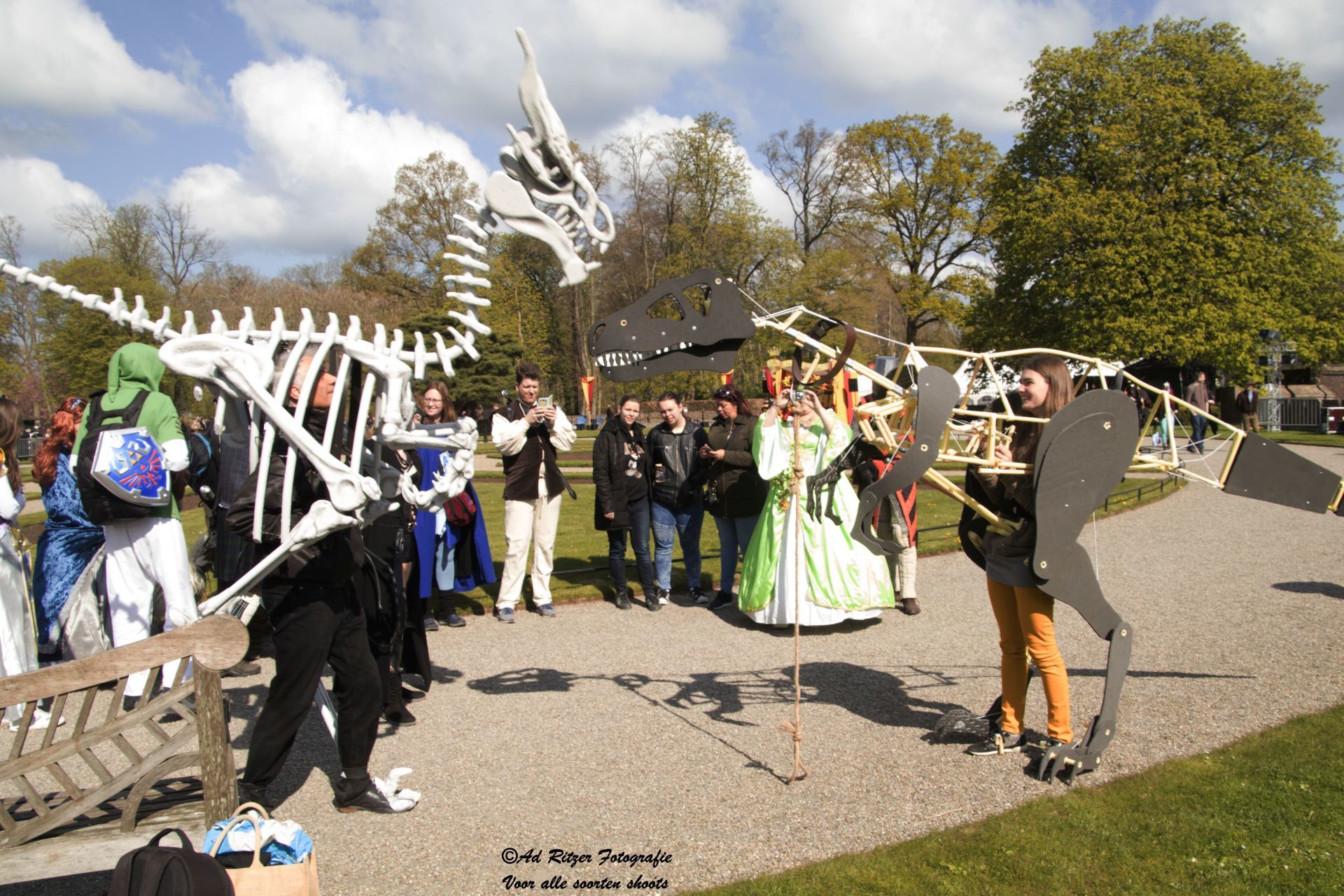 Mensen in kleurrijke kostuums op een openluchtfestival, met boven hun hoofden een wit skeletvormig drakenbeeld.