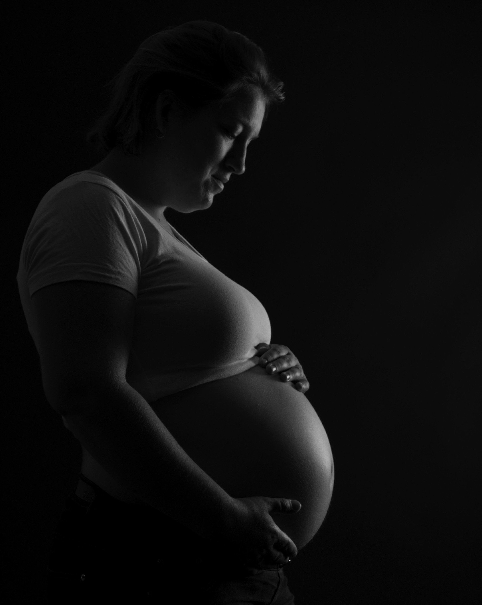 Silhouet van een zwangere vrouw in profiel, die zachtjes haar buik vasthoudt tegen een donkere achtergrond.