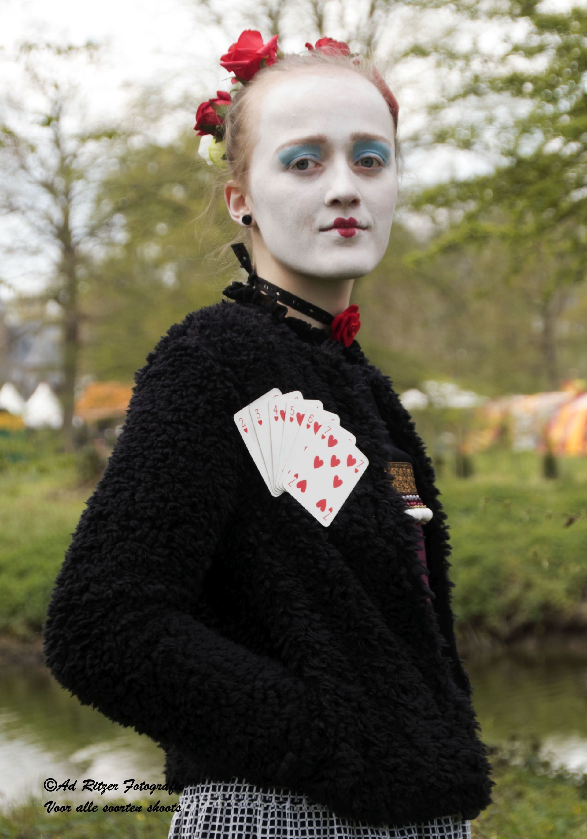 Persoon in zwarte trui met speelkaarten op de borst, witte gezichtsmake-up en rode bloemen in het haar, buiten.