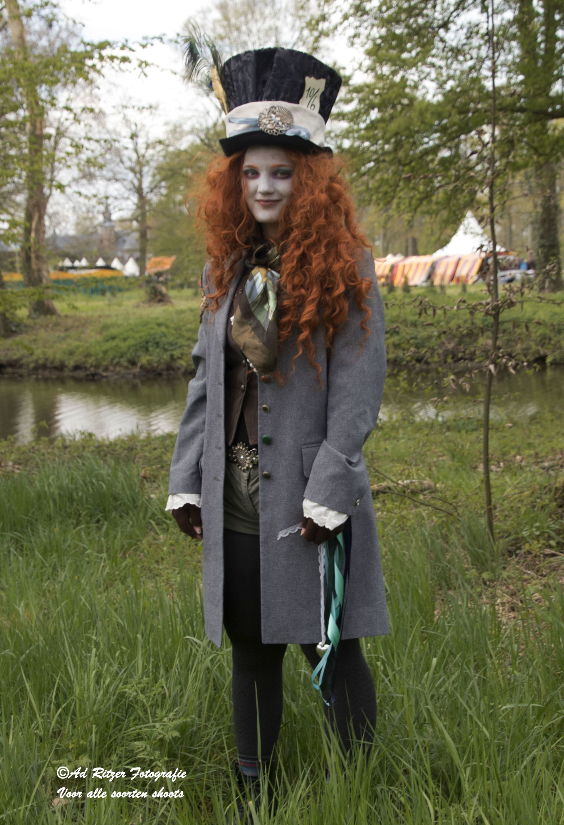Een persoon in een grijze jas en een hoge zwarte hoed staat bij een vijver in een grasrijk park.