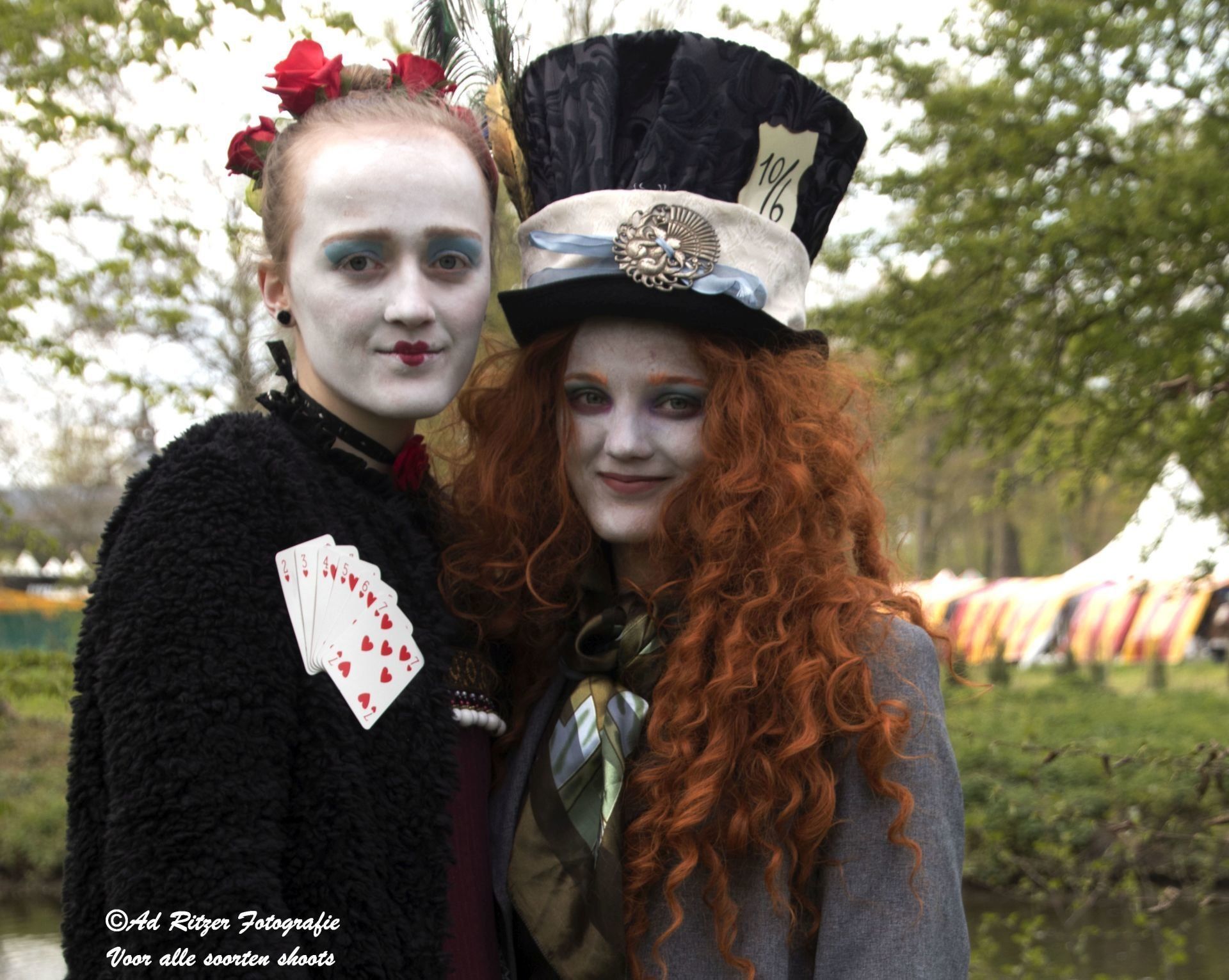 Twee verklede personen in een park, de een met speelkaarten in de hand, de ander met een hoge hoed op, beiden lachend naar de camera.