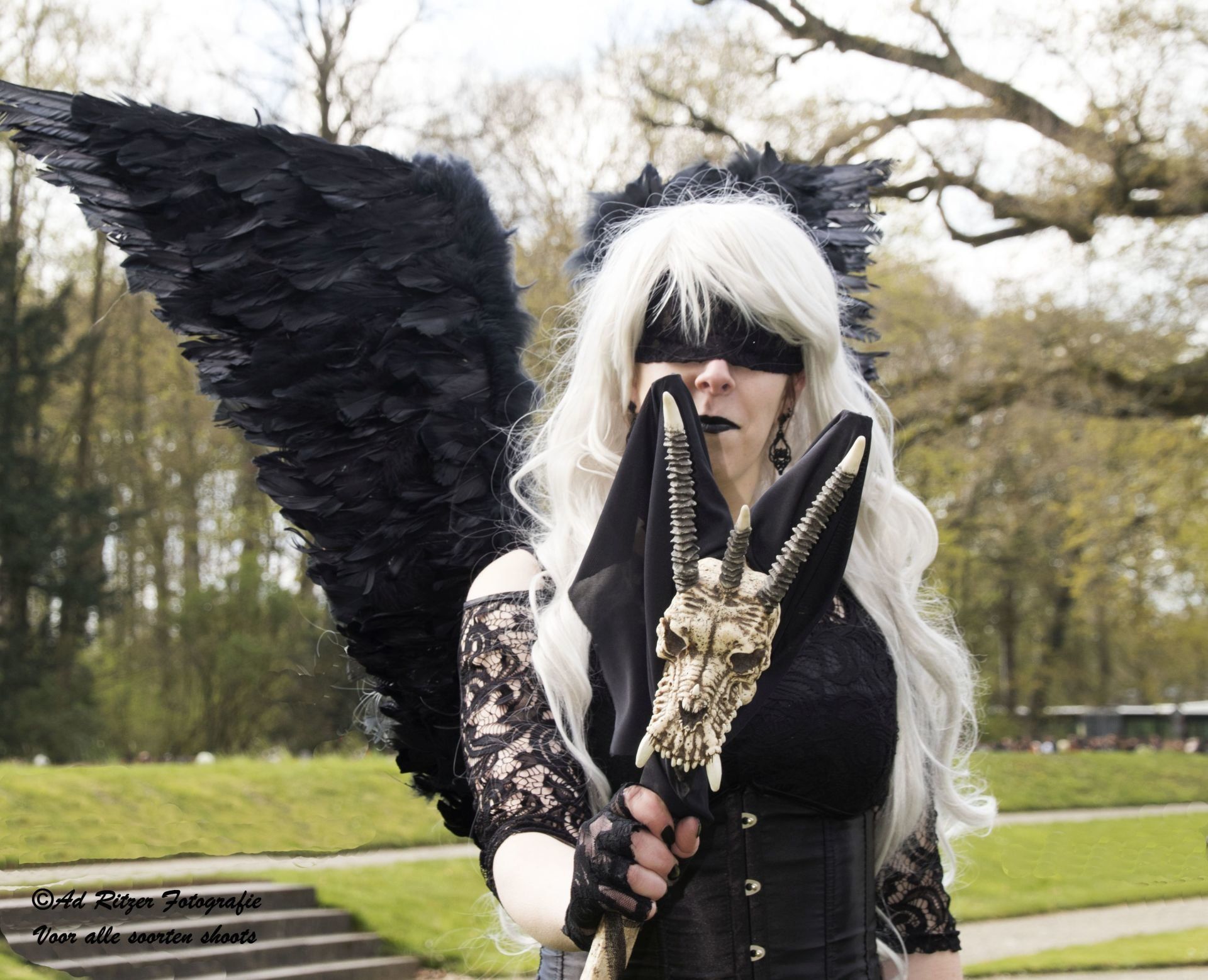 Een persoon in kostuum met zwarte vleugels en een witte pruik, die een staf vasthoudt in een park.
