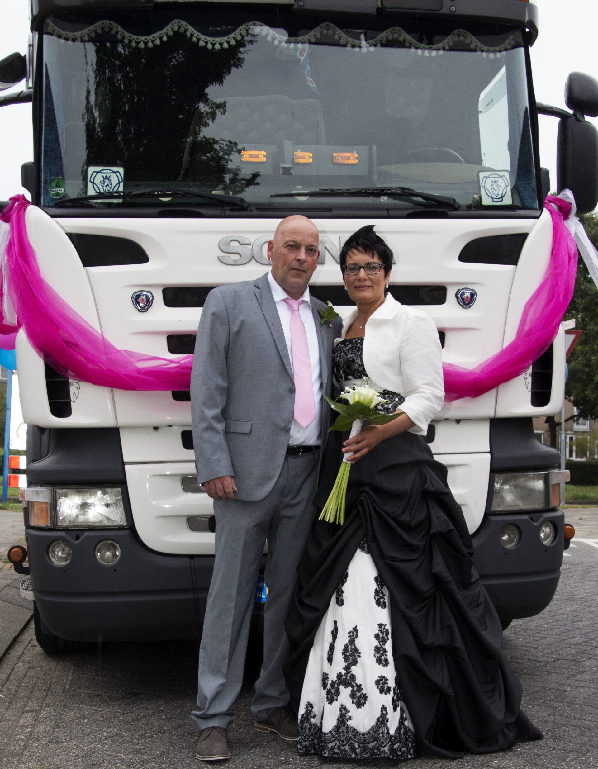 Een stel poseert in galakleding voor een versierde witte vrachtwagen met roze linten.