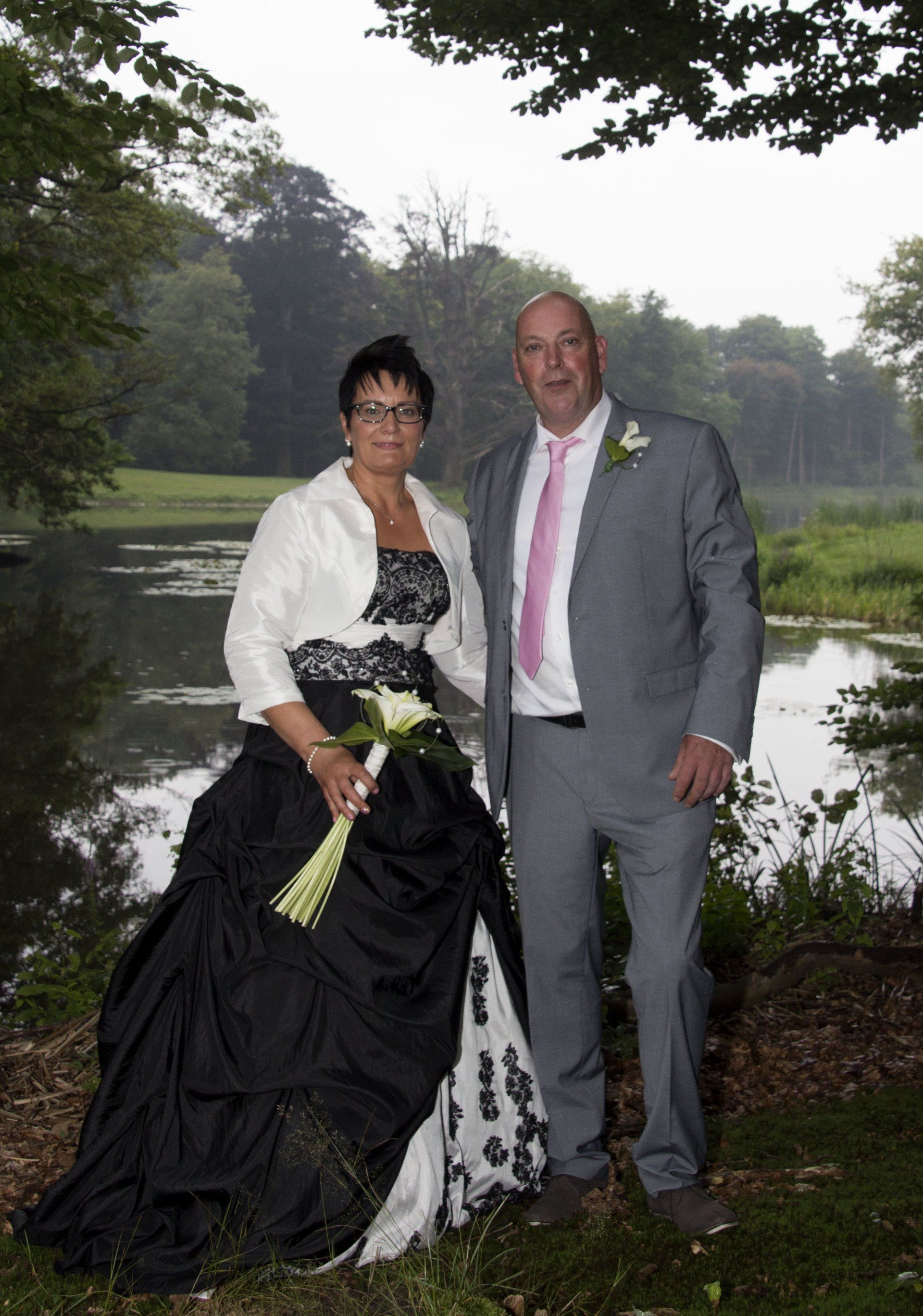 Een stel in formele kleding poseert bij een vijver in een bosrijke tuin.