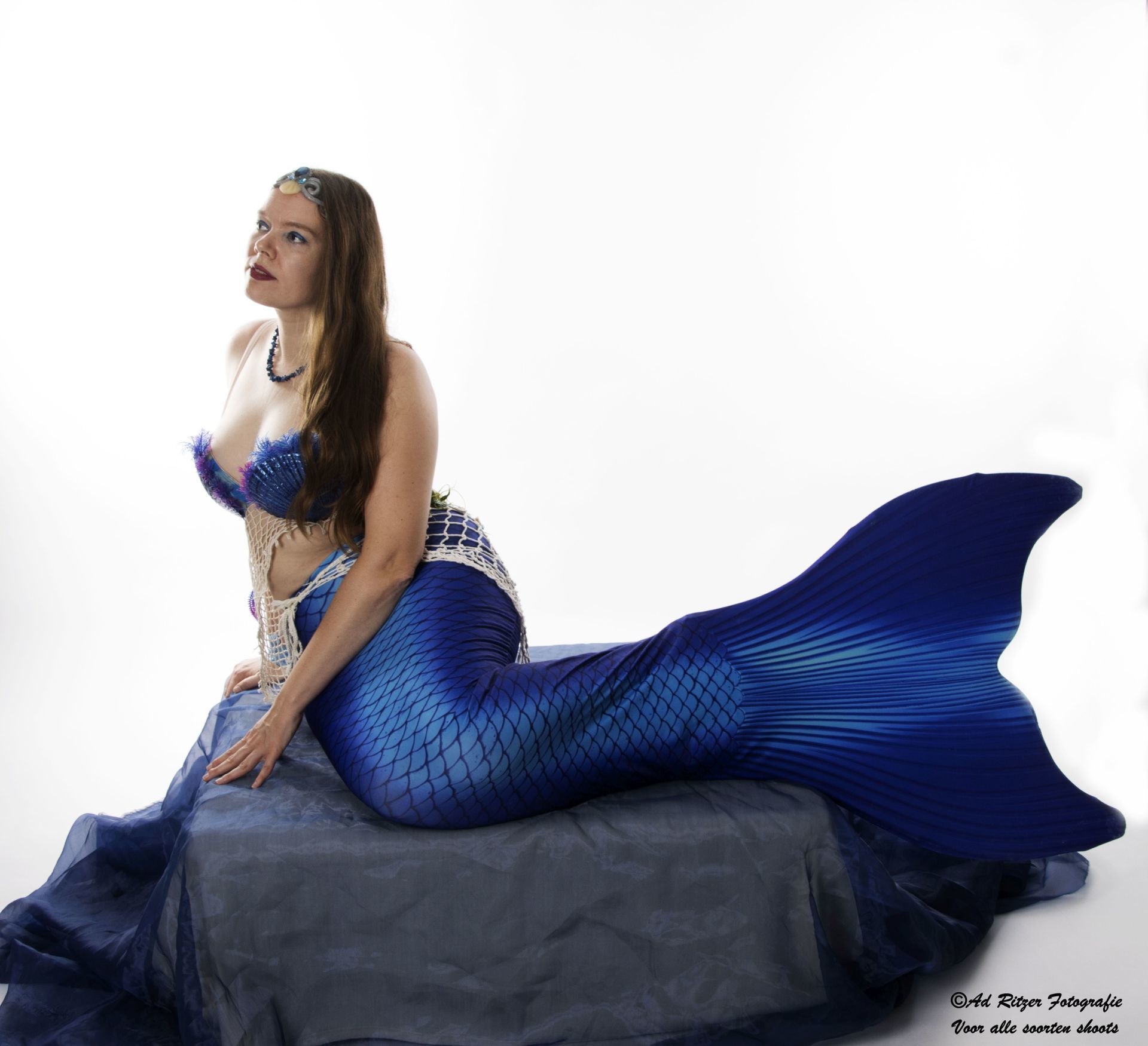 Vrouw met een blauwe zeemeerminnenstaart poseert op een rots tegen een witte achtergrond.