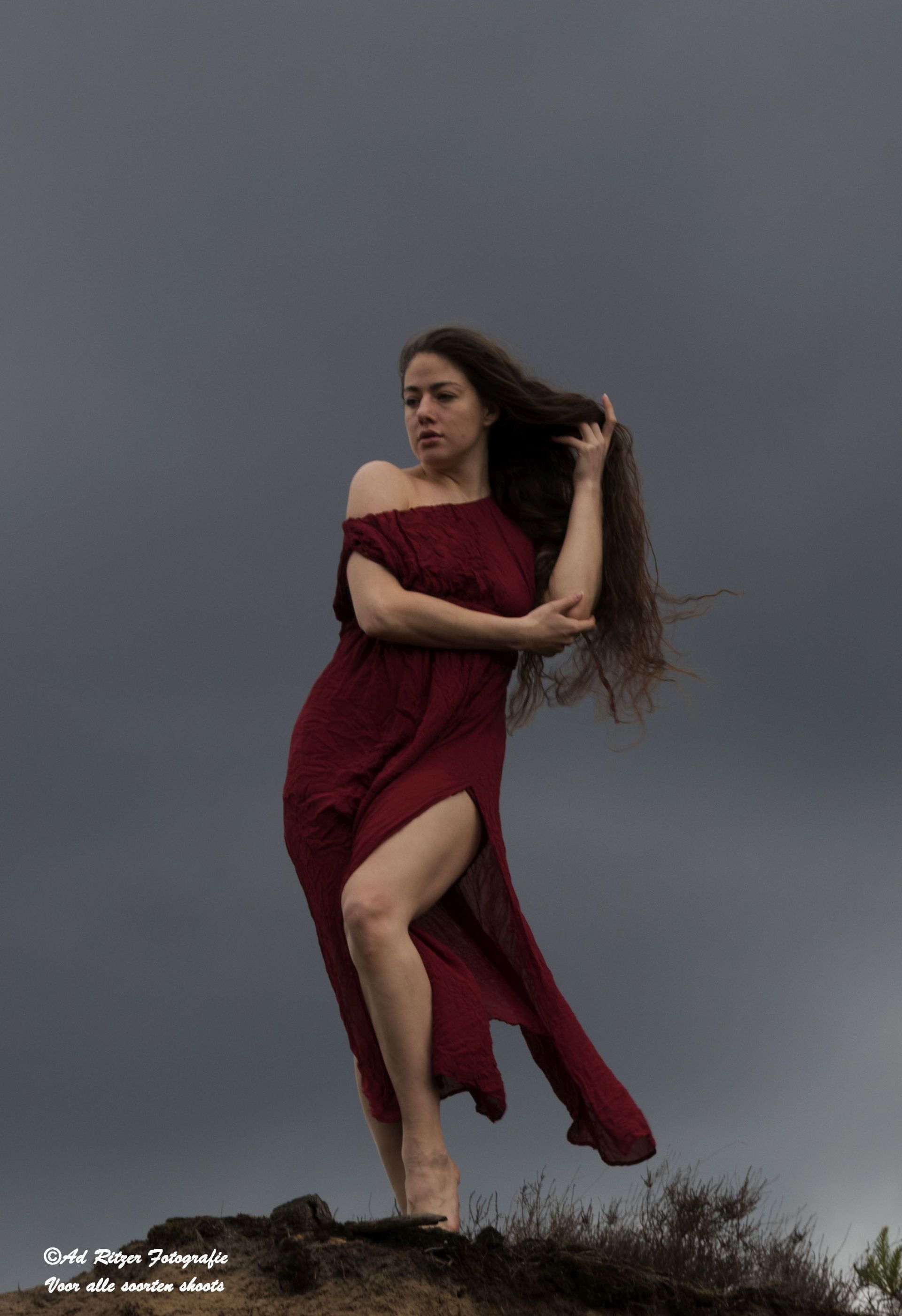 Vrouw in een zwierige rode jurk staat op rotsen in de wind tegen een grijze hemel.