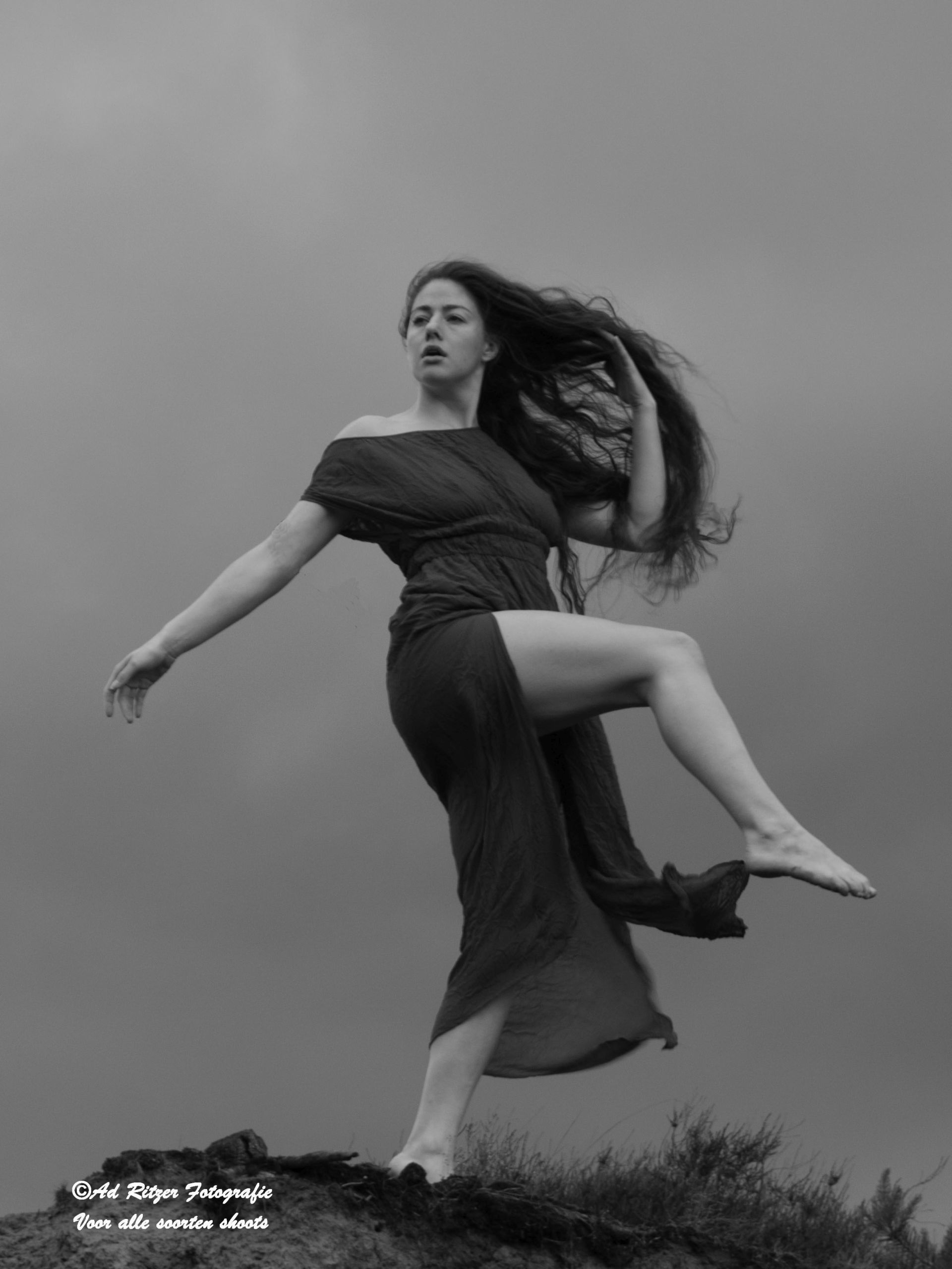 Vrouw in een zwierige jurk poseert buiten op een rotsachtige heuvel, haar haar wapperend in de wind.