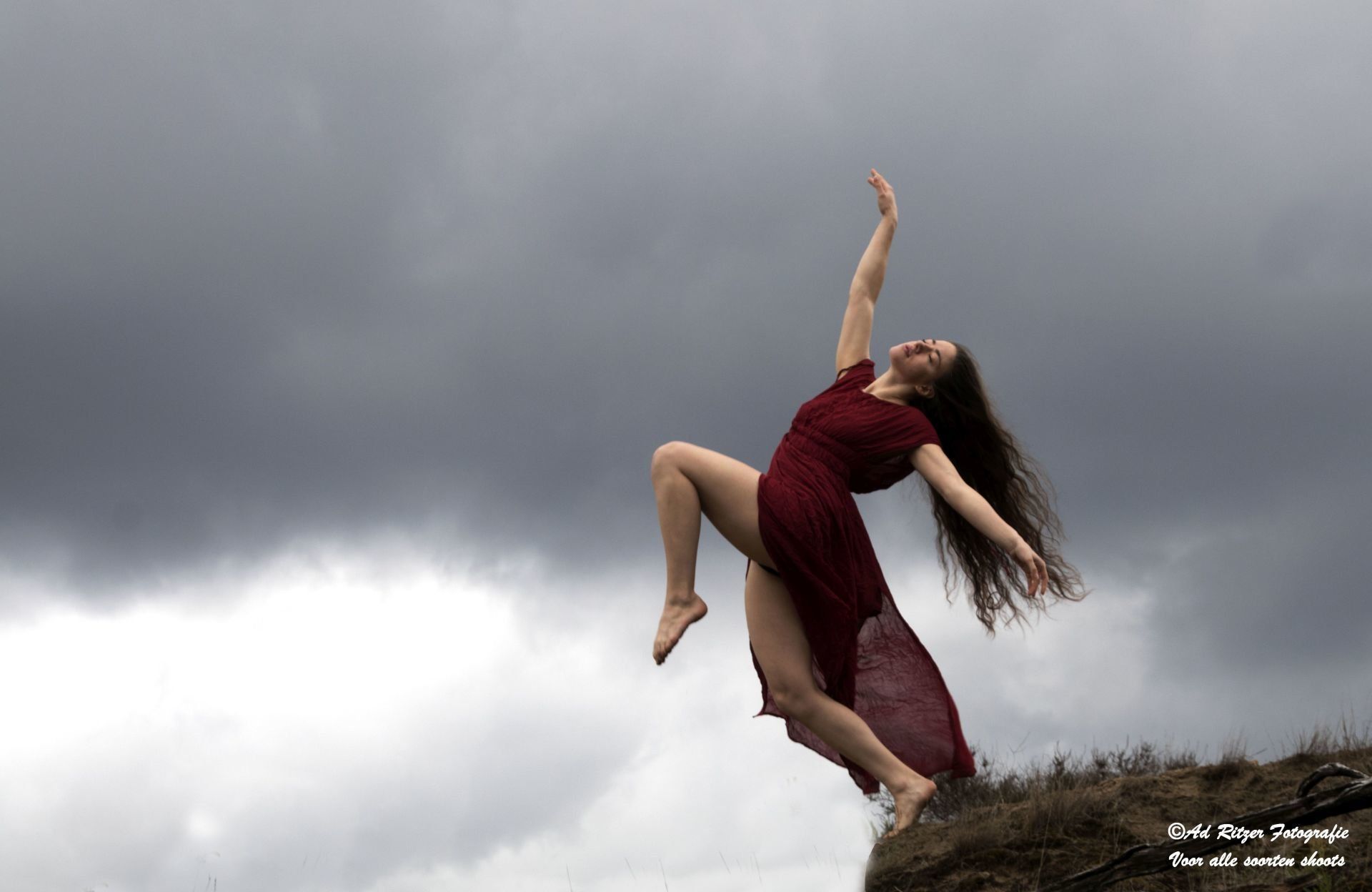 Vrouw in een rode jurk springt van een klif onder donkere stormwolken.