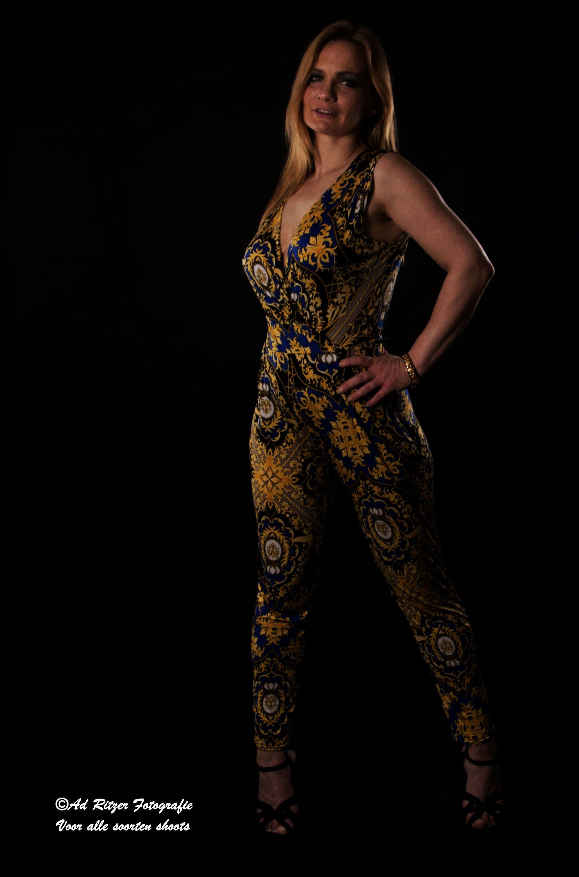 Vrouw in een goud-zwart gestreepte jumpsuit poseert tegen een zwarte achtergrond.