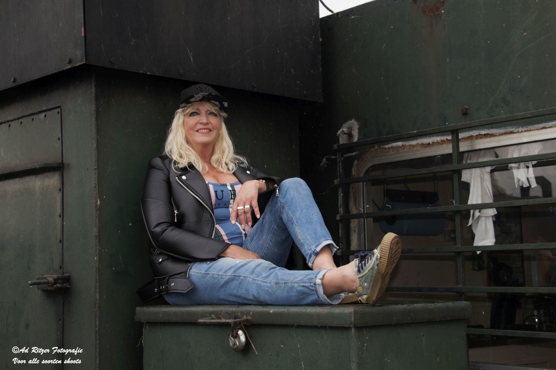 Vrouw zit op een donkergroene richel, glimlachend, gekleed in een zwarte jas, spijkerbroek en hoed.