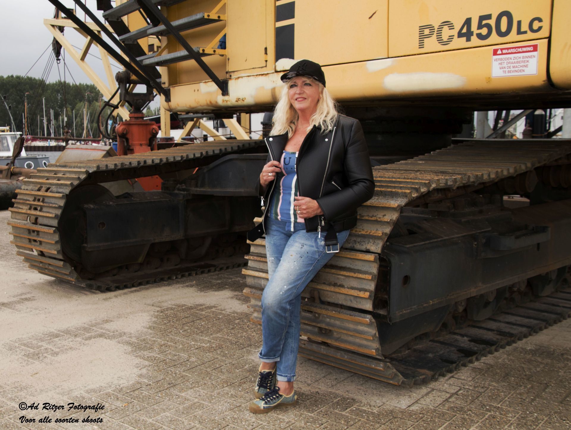 Vrouw staat naast een grote gele graafmachine met rupsbanden op een bouwterrein.
