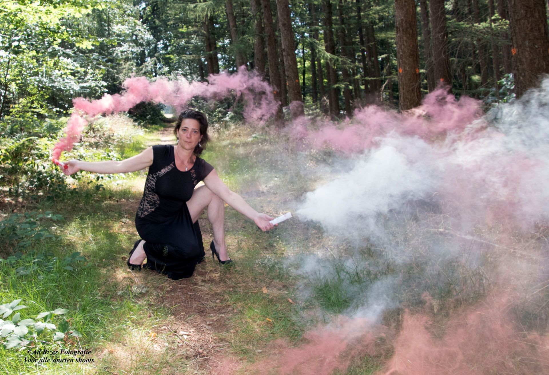 Een persoon knielt in een bos en houdt roze rookfakkels vast die wolken van roze en witte rook produceren.