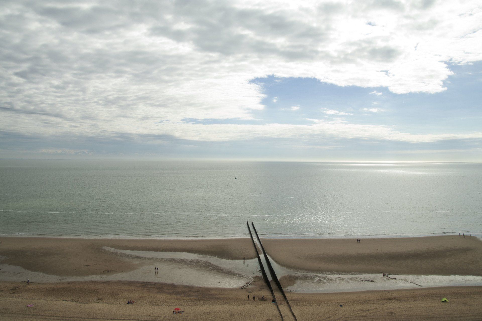 Een breed strand onder een bewolkte hemel, met een kalme oceaan en twee scheefstaande palen op de voorgrond.