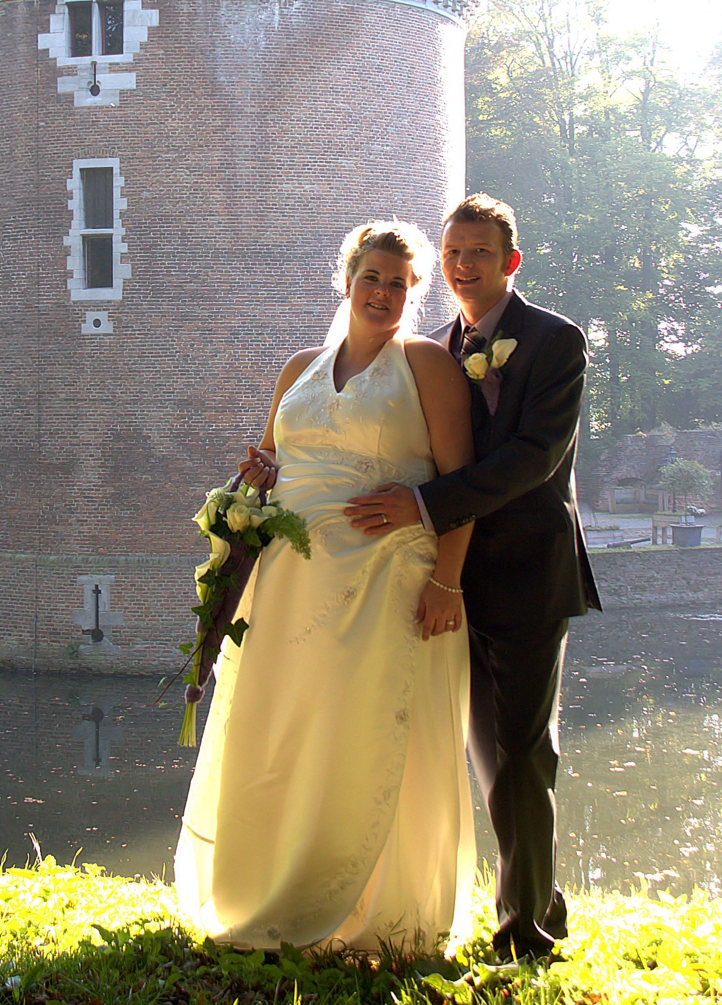 Bruid en bruidegom poseren buiten bij een bakstenen toren, met zonlicht en water op de achtergrond.