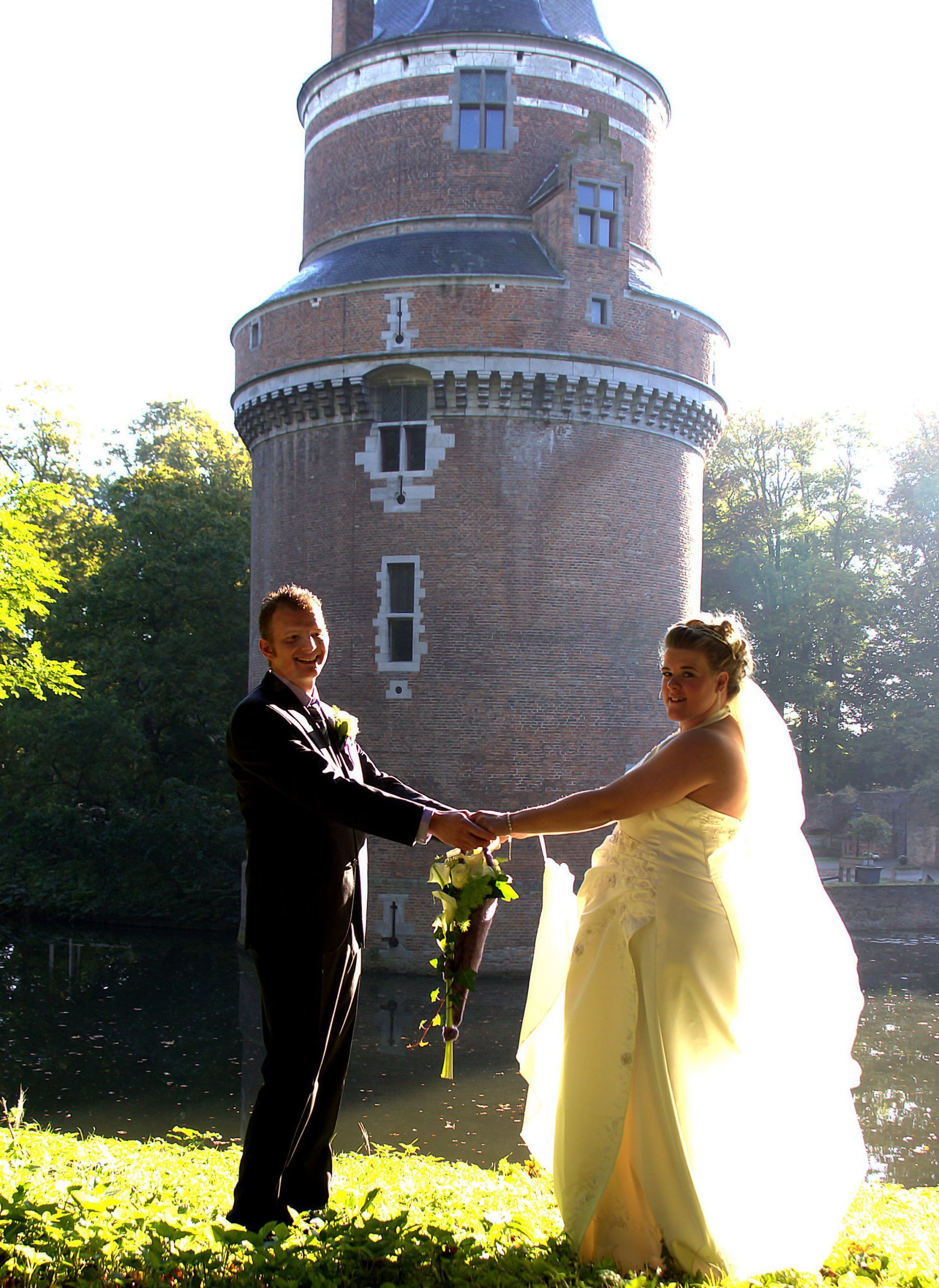 Een bruidspaar, in trouwkleding, loopt hand in hand voor een bakstenen toren op een zonnig gazon.