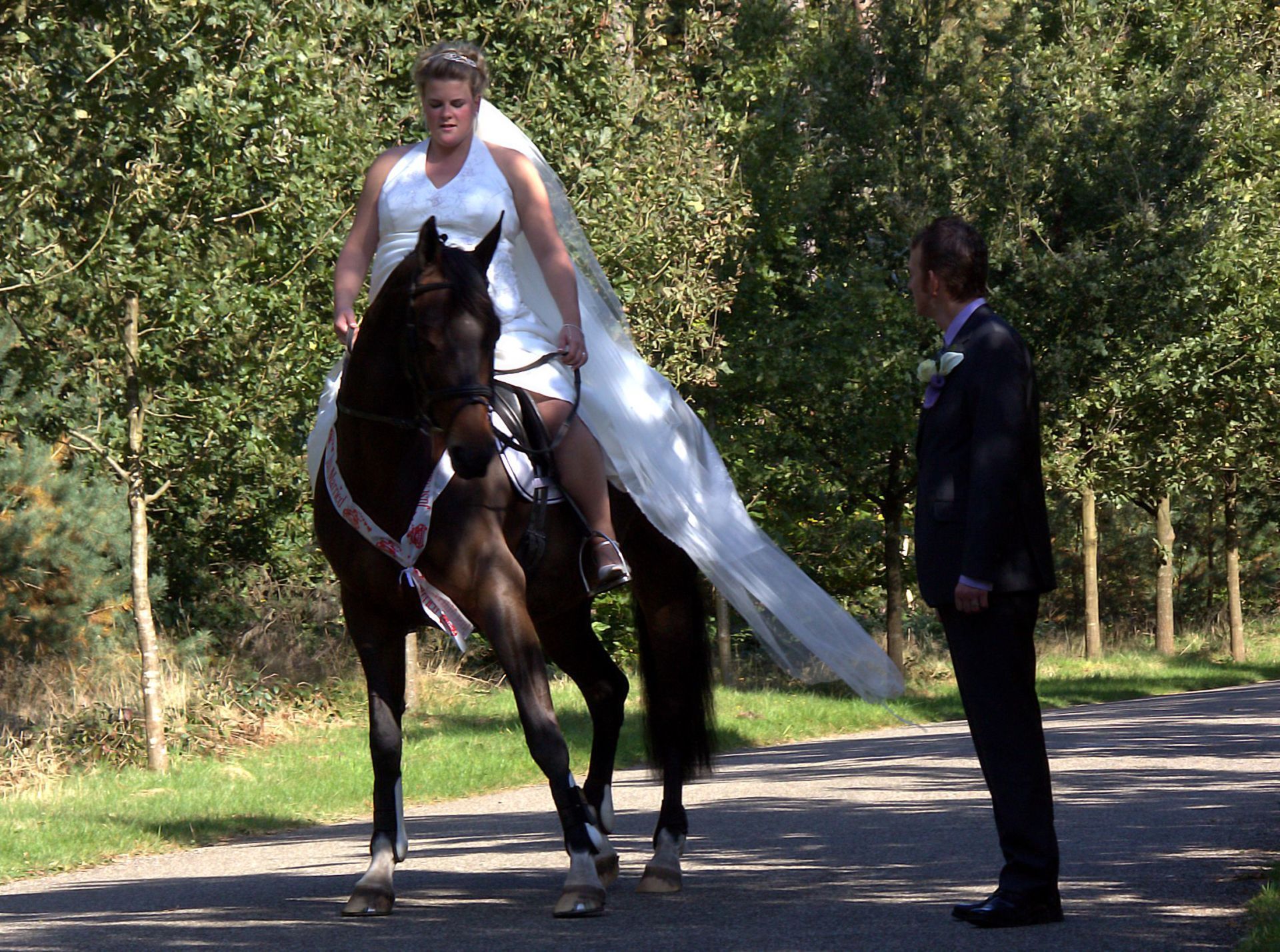 Bruid in witte jurk te paard naast een man in een donker pak op een met bomen omzoomd pad.