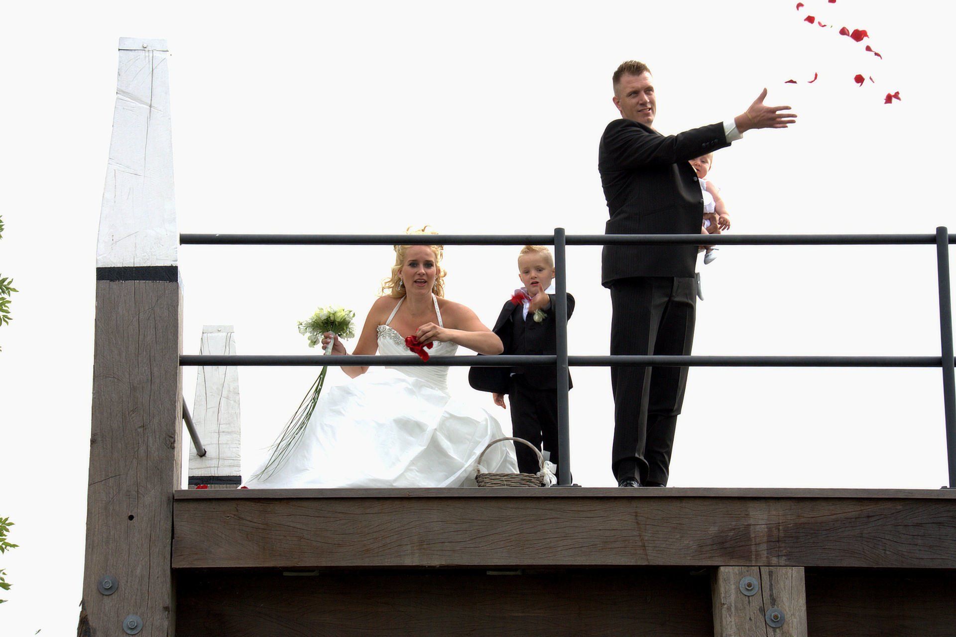 Een man in een zwart pak gooit rode bloemblaadjes vanaf een balkon naar beneden, terwijl twee mensen toekijken.