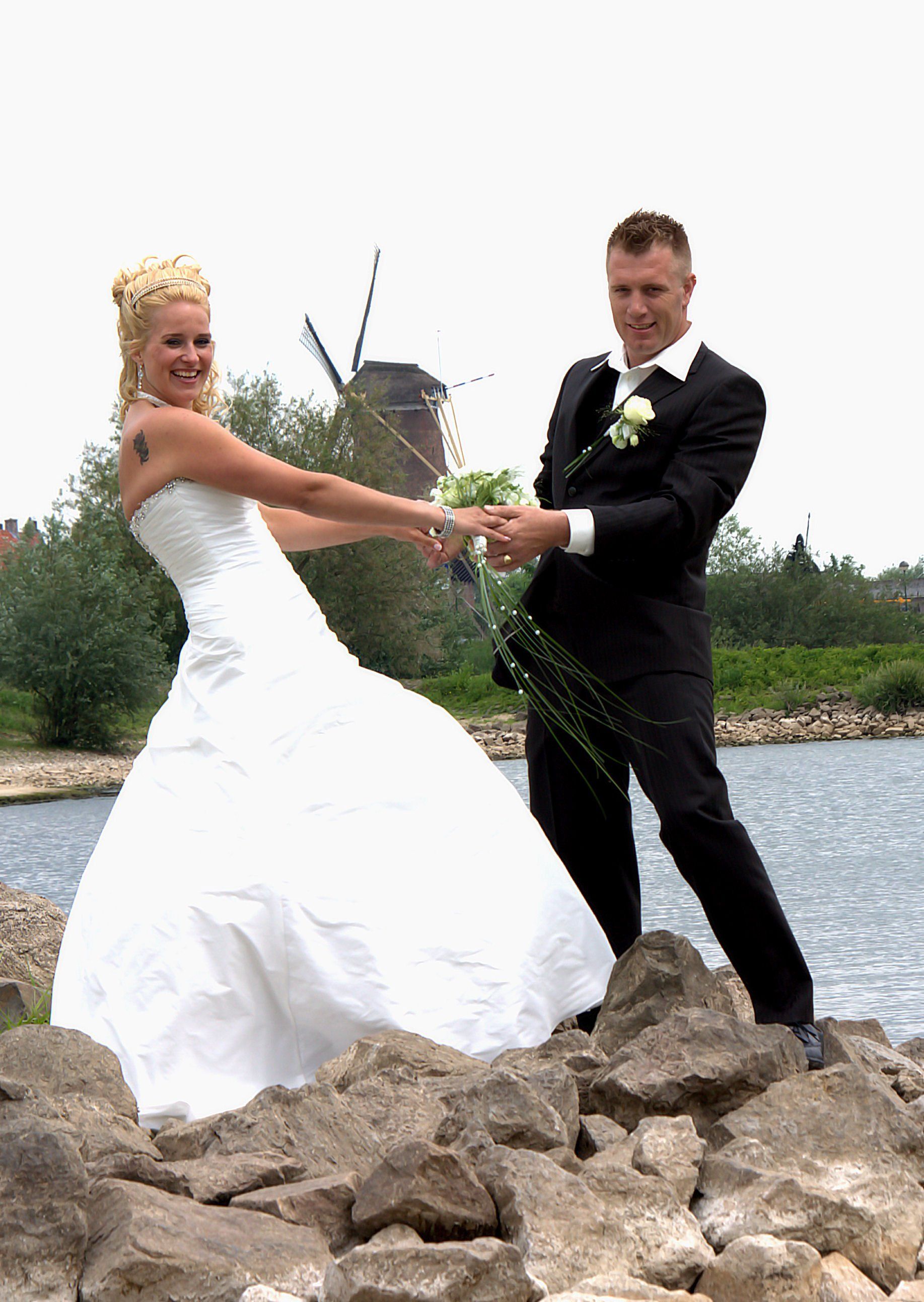 Bruid en bruidegom in trouwkleding, hand in hand op rotsen aan het water, met een windmolen op de achtergrond.