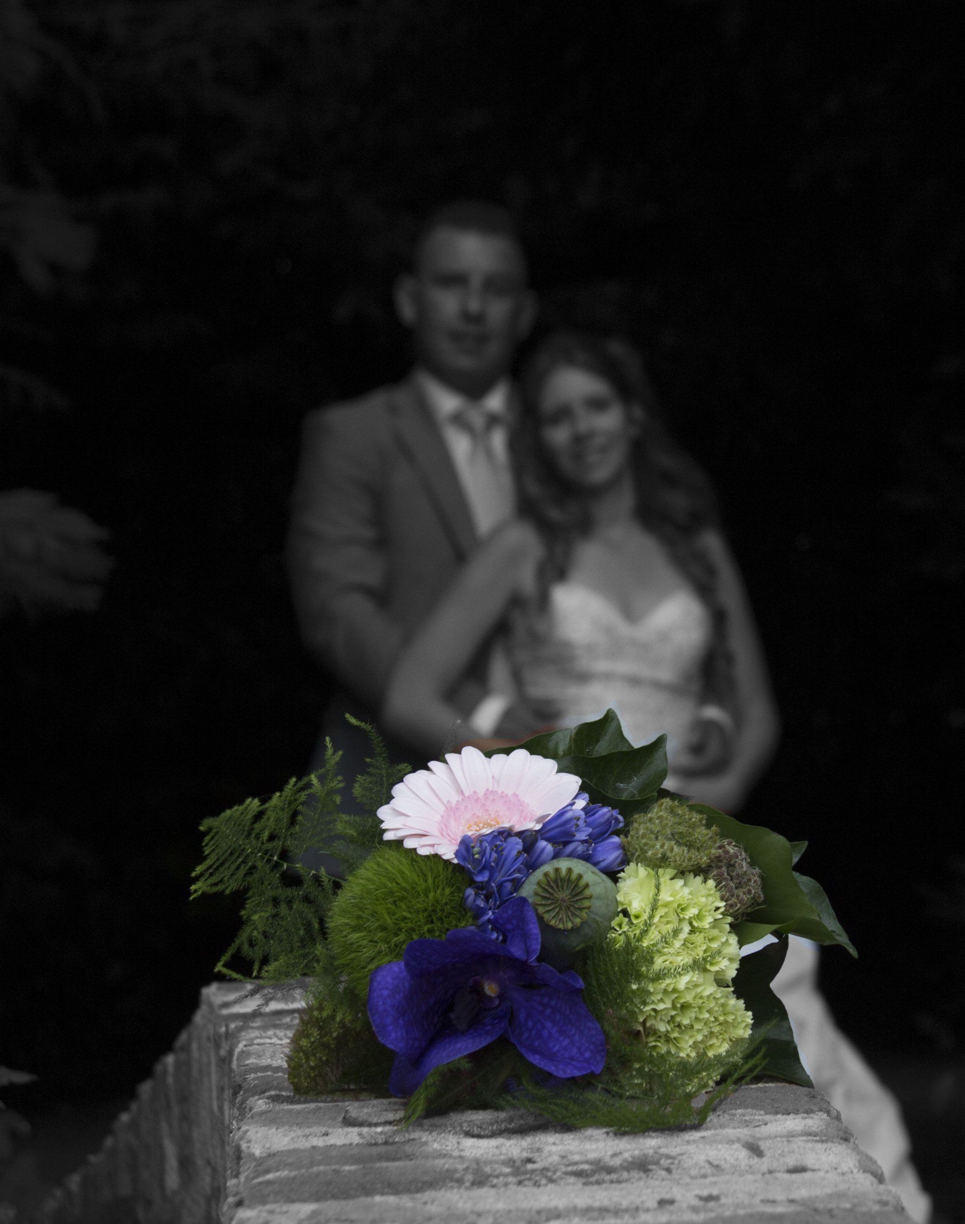 Bruidspaar achter een boeket paarse, witte en groene bloemen in een donkere omgeving.