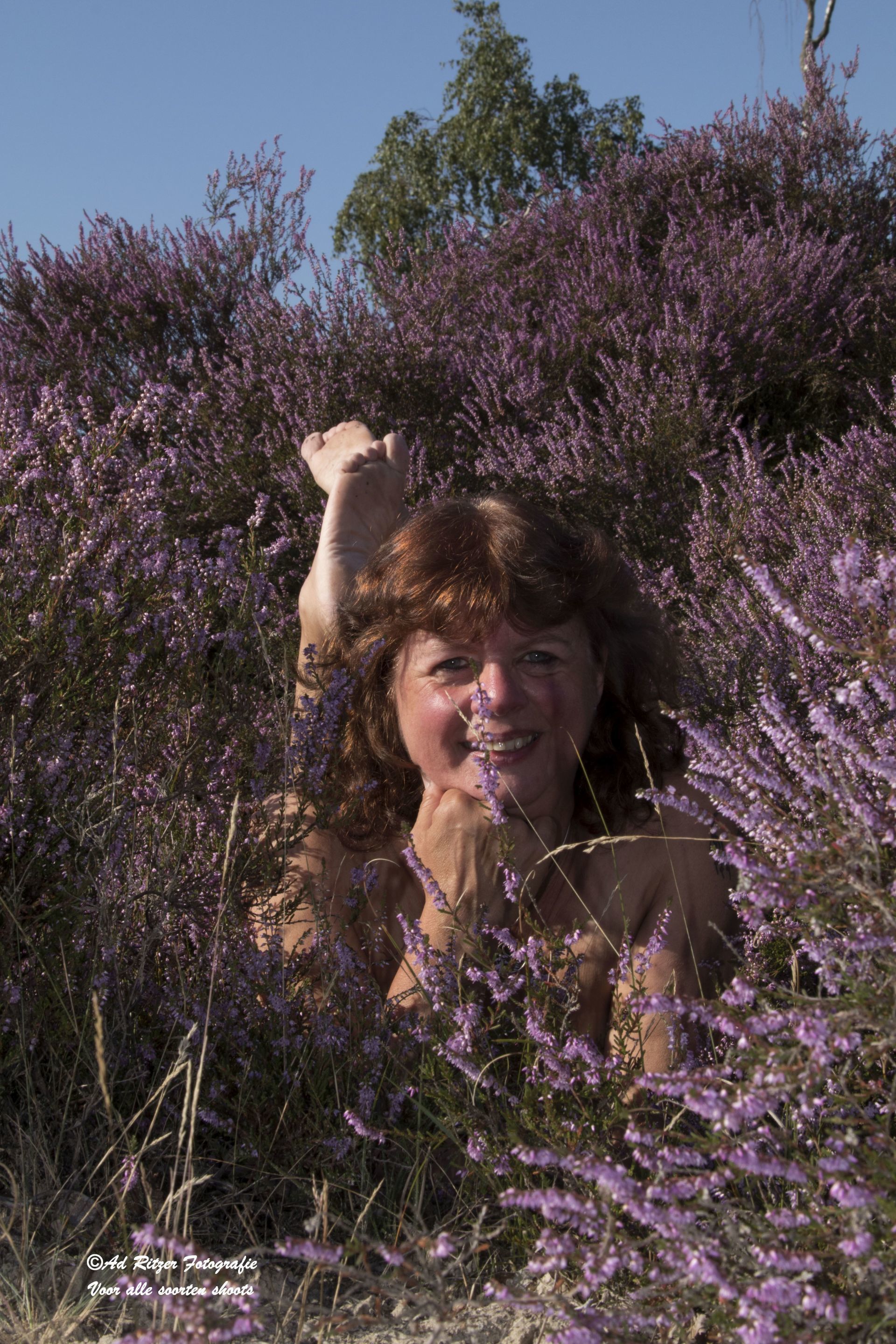 Een persoon lacht te midden van paarse bloemen, met een opgeheven arm in een zonovergoten veld.