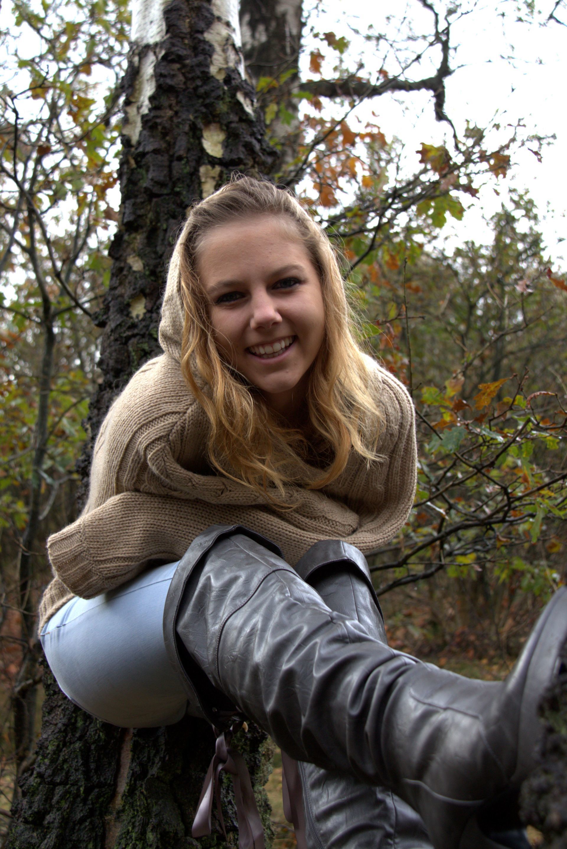 Een lachende vrouw in een beige trui en grijze laarzen zit op een boom in een bosrijk gebied.