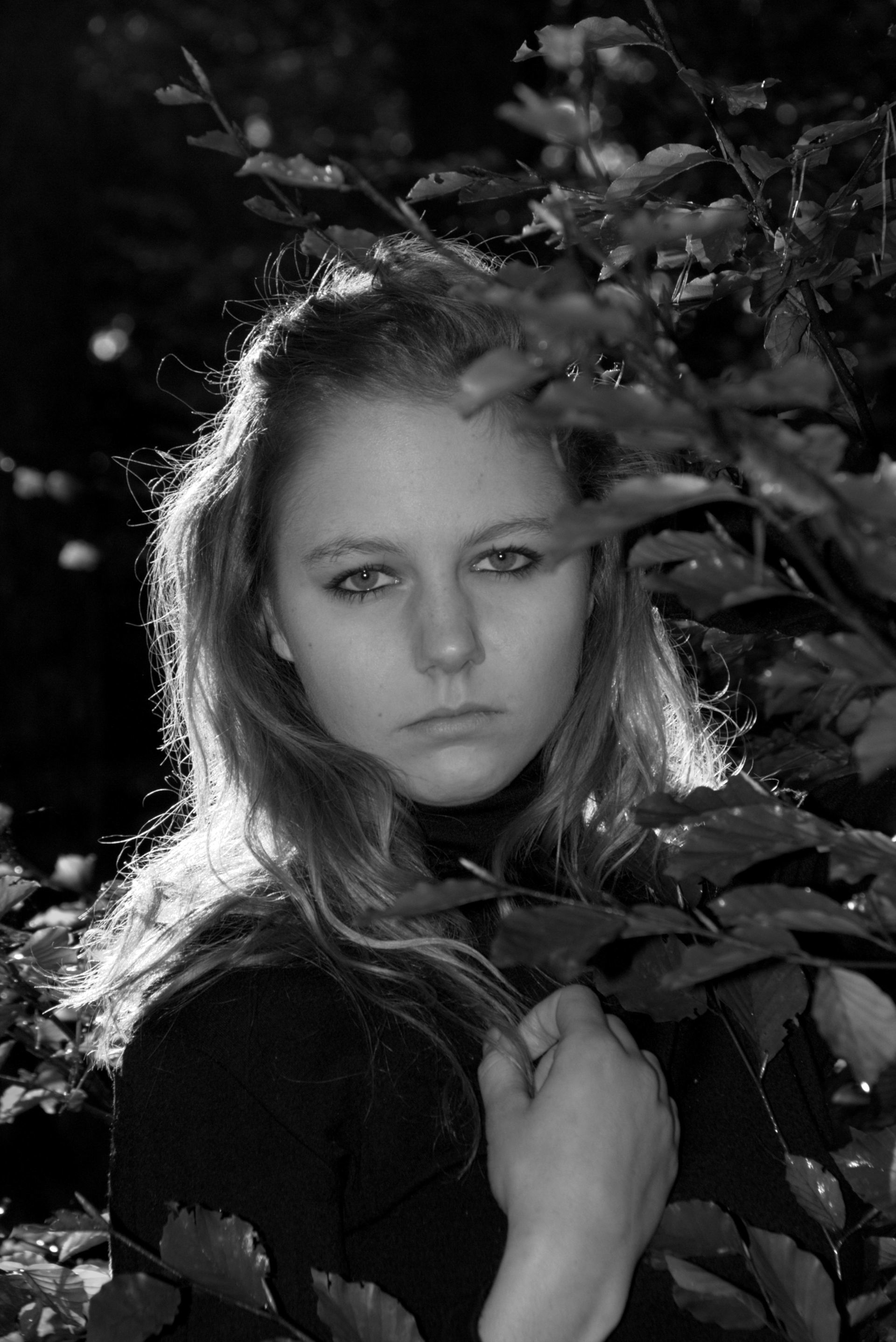Portret van een vrouw in donkere kleding, staand tussen bladerrijke takken, die in de camera kijkt.