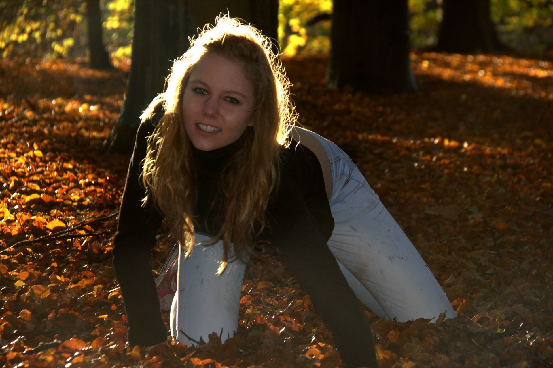 Een persoon in een witte jas hurkt buiten in het zonovergoten herfstblad en lacht naar de camera.