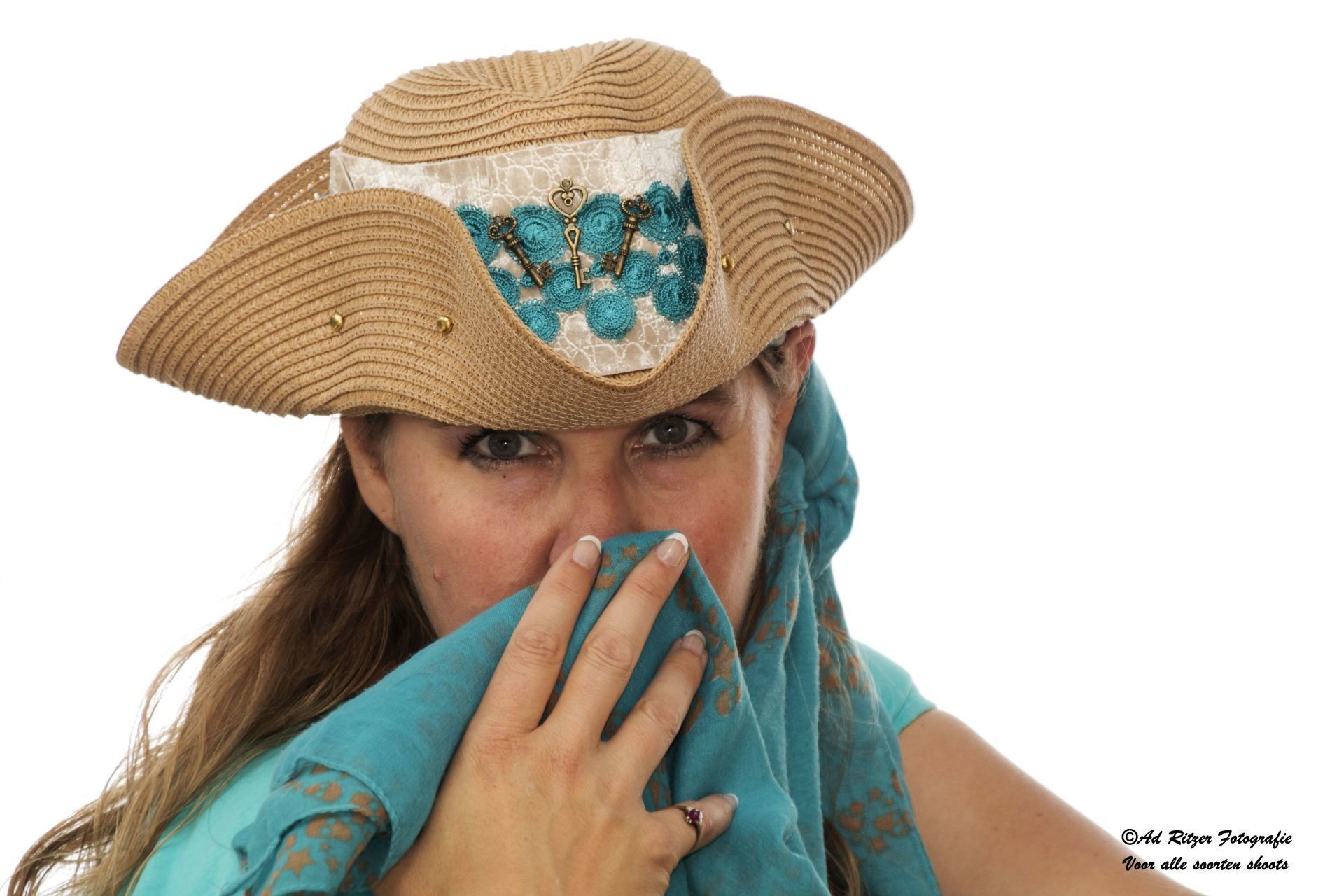 Een vrouw met een strohoed houdt een turquoise doek voor haar gezicht, waar haar ogen doorheen turen.