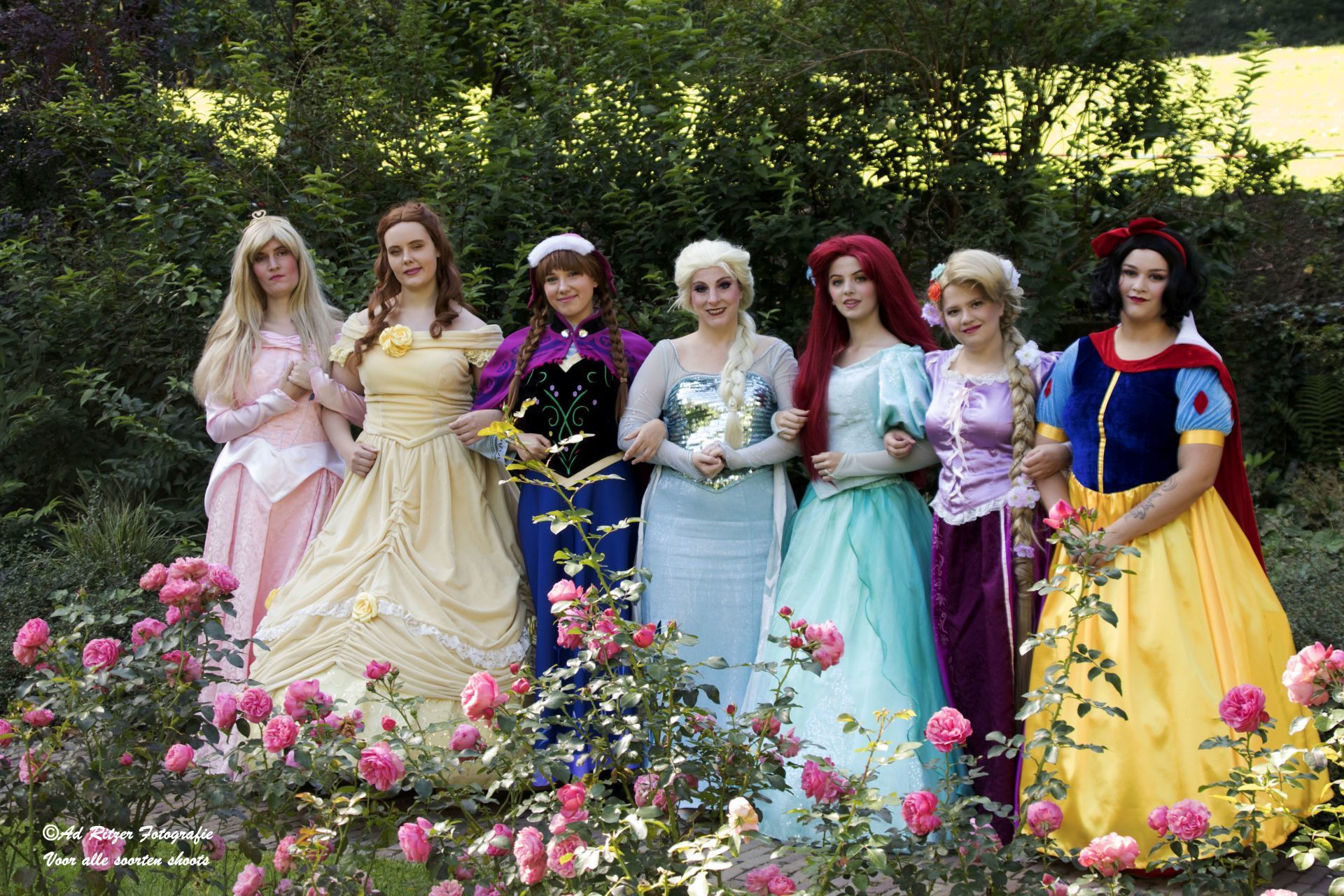 Zes mensen in kleurrijke prinsessenkostuums poseren tussen bloeiende rozen in een tuin.
