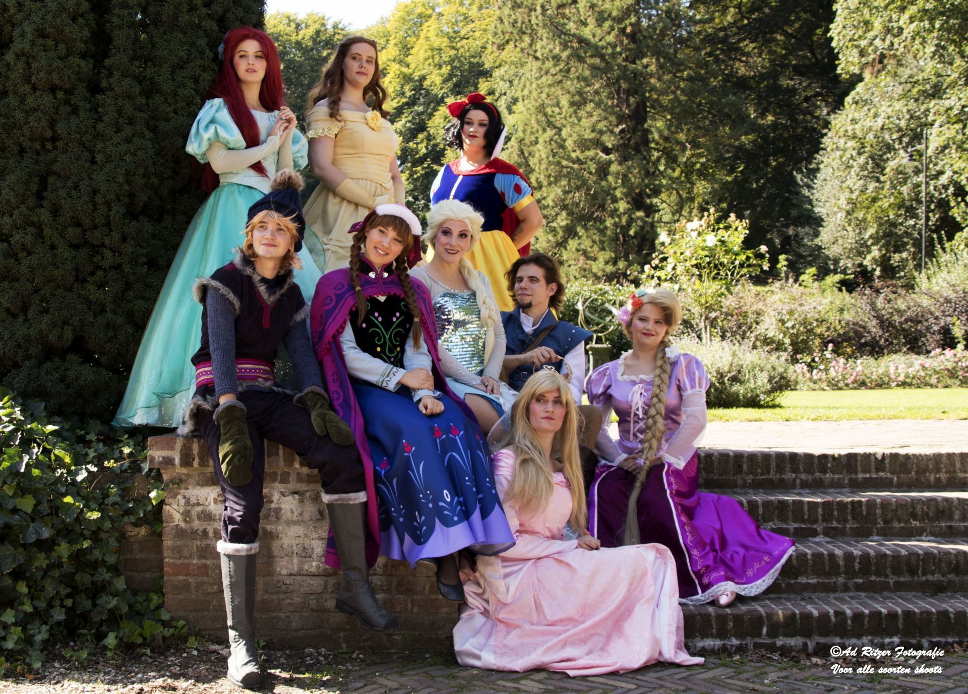 Een groep in kostuums poseert op stenen trappen in een zonovergoten tuin, met kleurrijke jurken en pakken in prinsessenstijl.