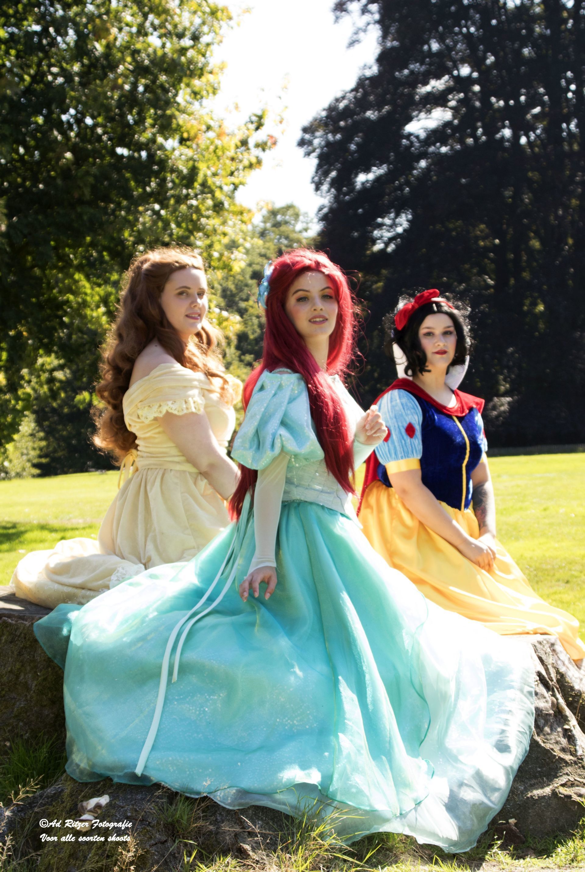 Drie vrouwen in kleurrijke prinsessenjurken zitten in de zon op een grasveld.