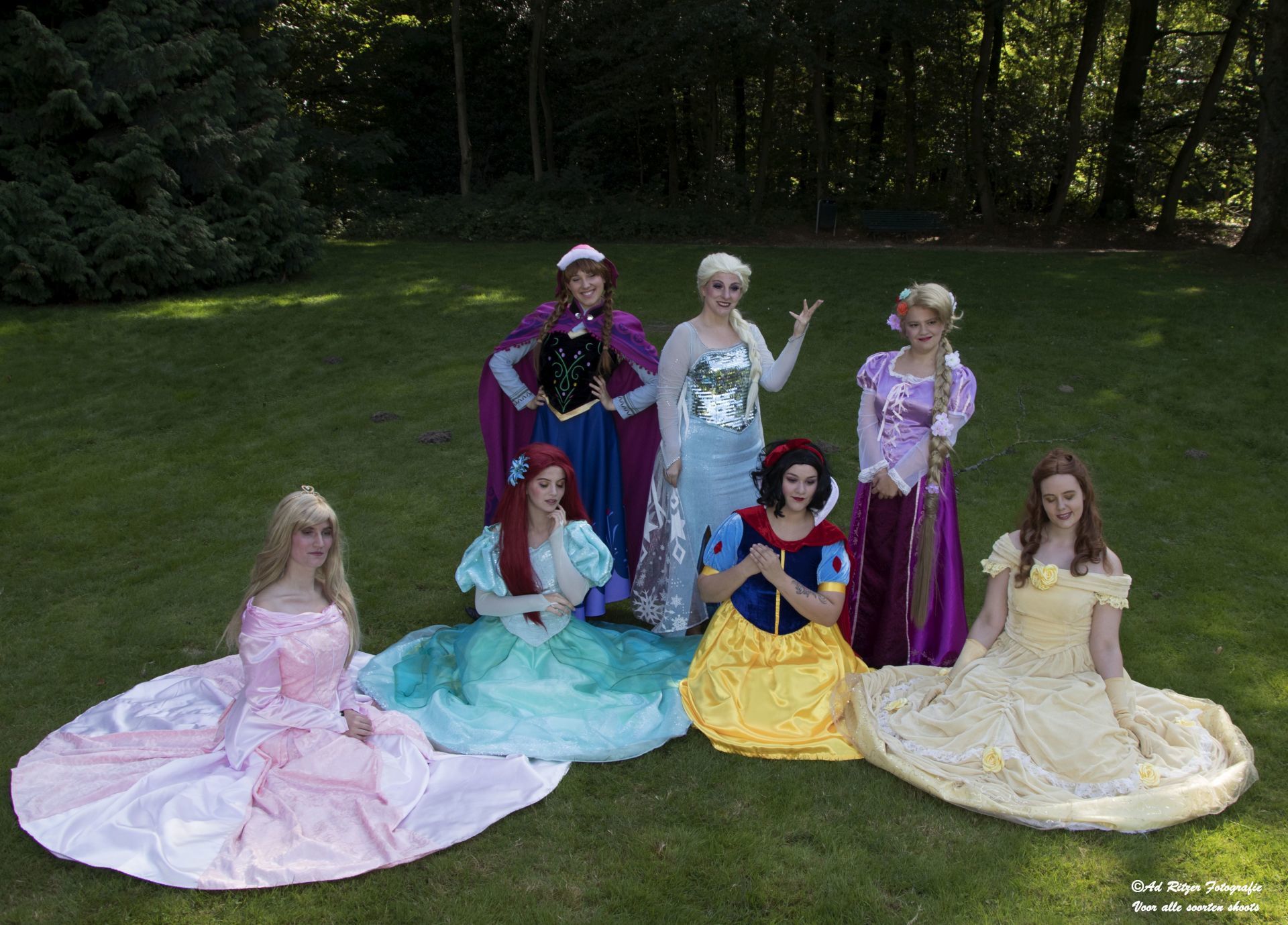 Zeven kinderen in prinsessenjurken poseren buiten op het gras in een bosrijk park.