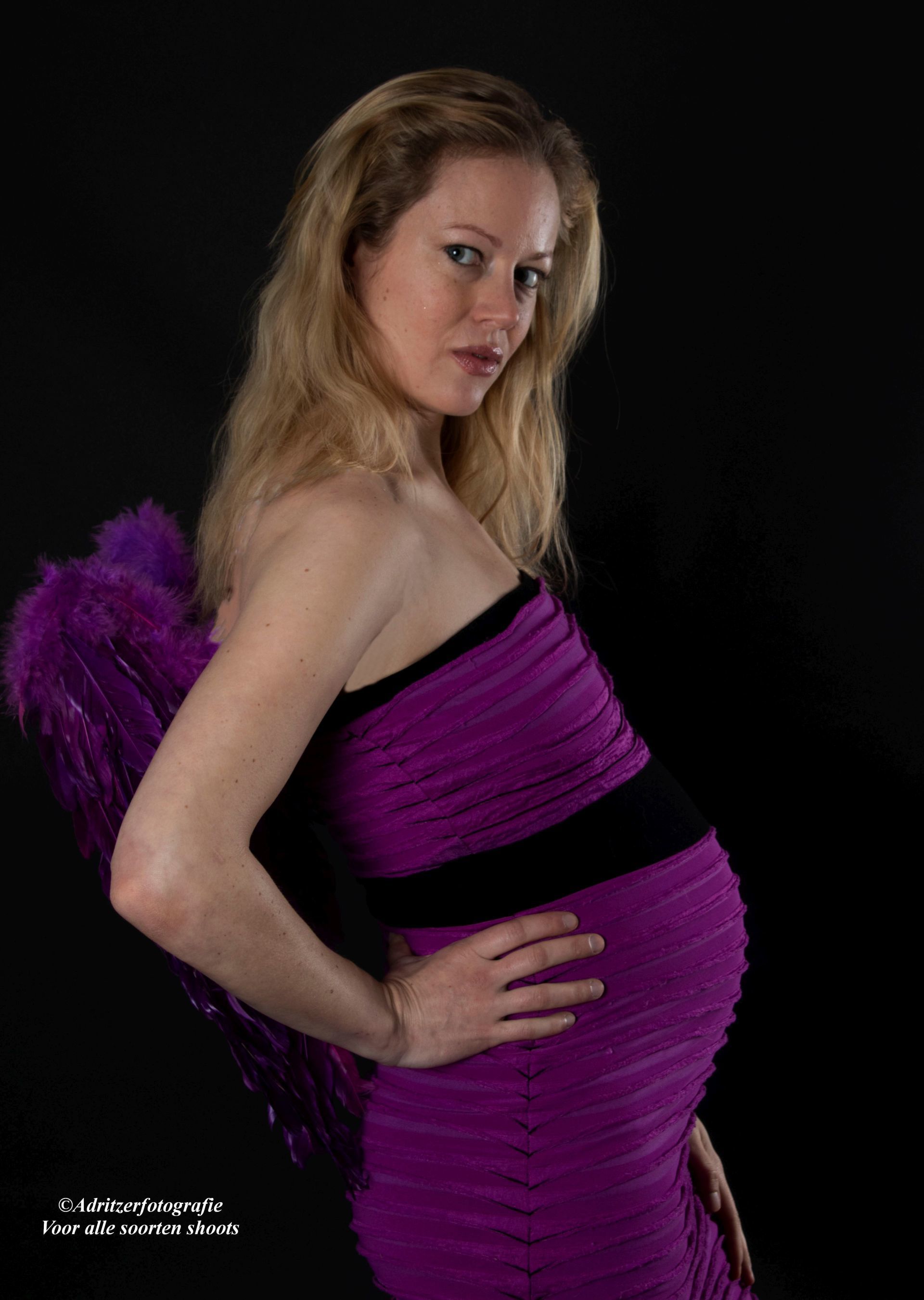 Vrouw in een paarse jurk poseert tegen een zwarte achtergrond en kijkt over haar schouder.