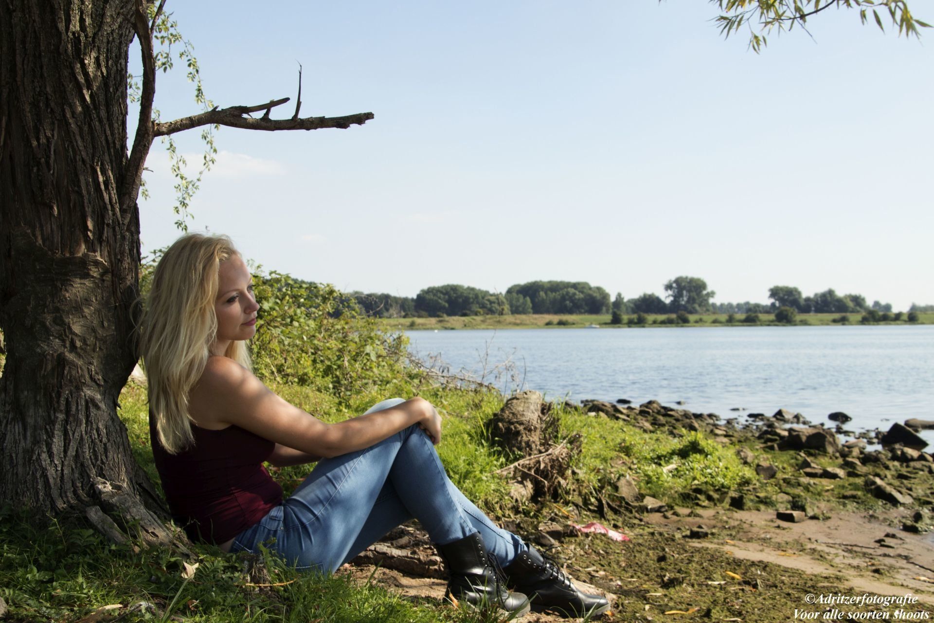 Een persoon zit onder een boom aan de oever van een meer en kijkt uit over het water.