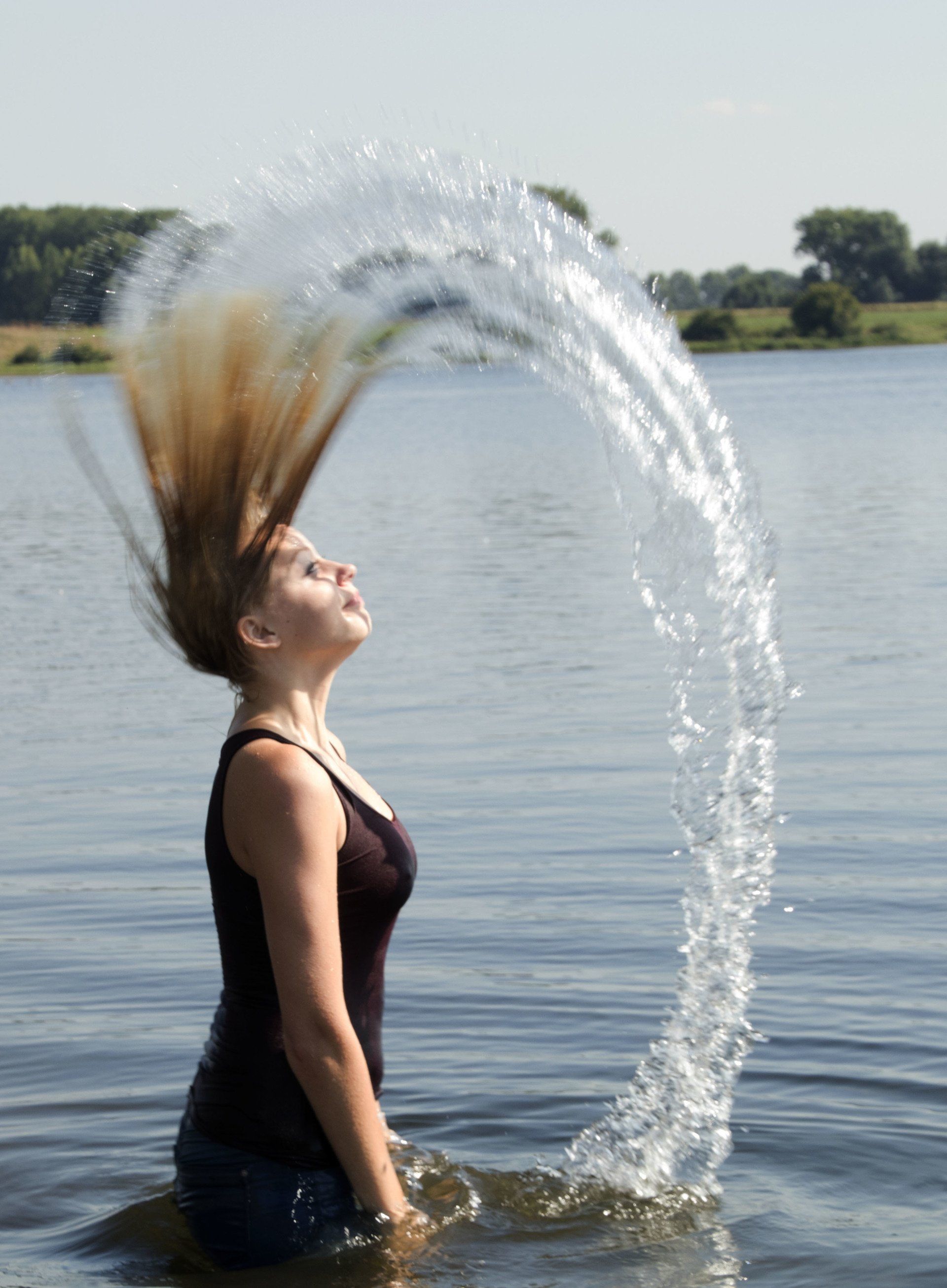 Een persoon in een meer gooit water in een boog vanuit zijn natte haar, met bomen zichtbaar op de achtergrond.