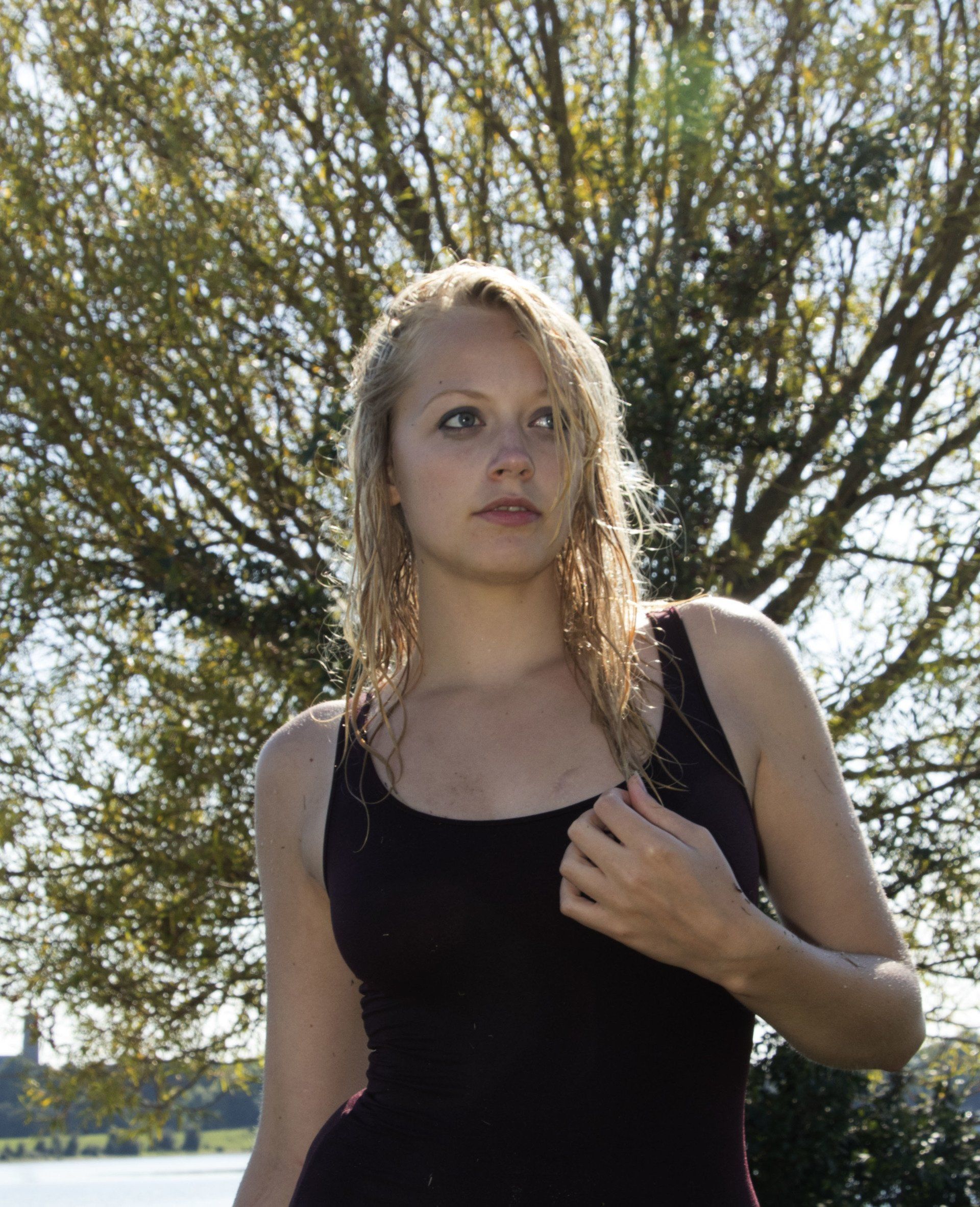 Een blond persoon in een zwarte top staat buiten voor een boom in de zon.
