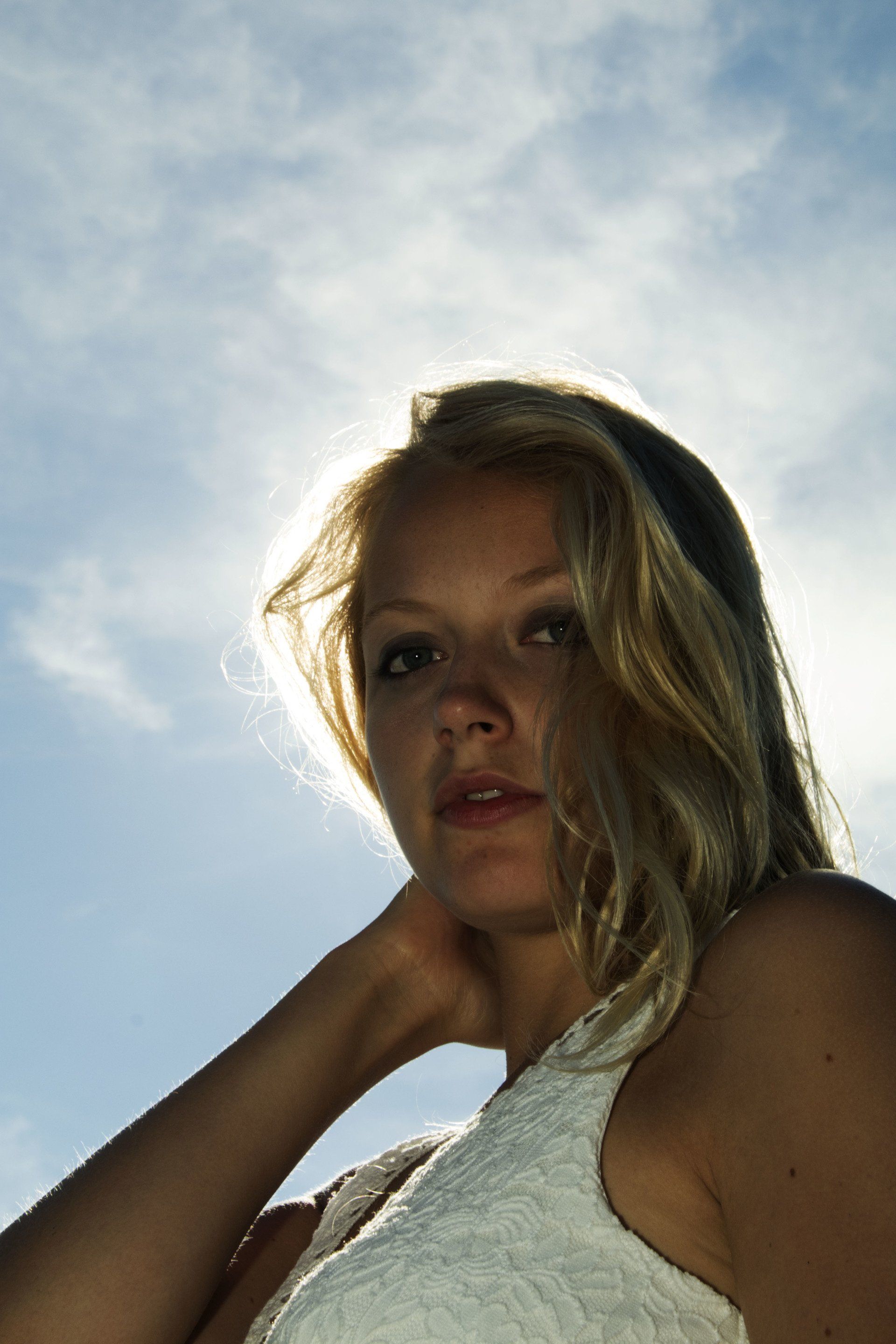 Blonde vrouw in een witte top kijkt in de camera tegen een heldere, bewolkte hemel.