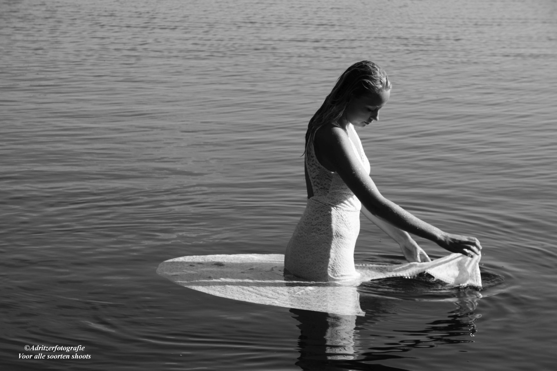 Vrouw in een witte jurk staat in ondiep water, kijkt naar beneden en houdt de stof vast.