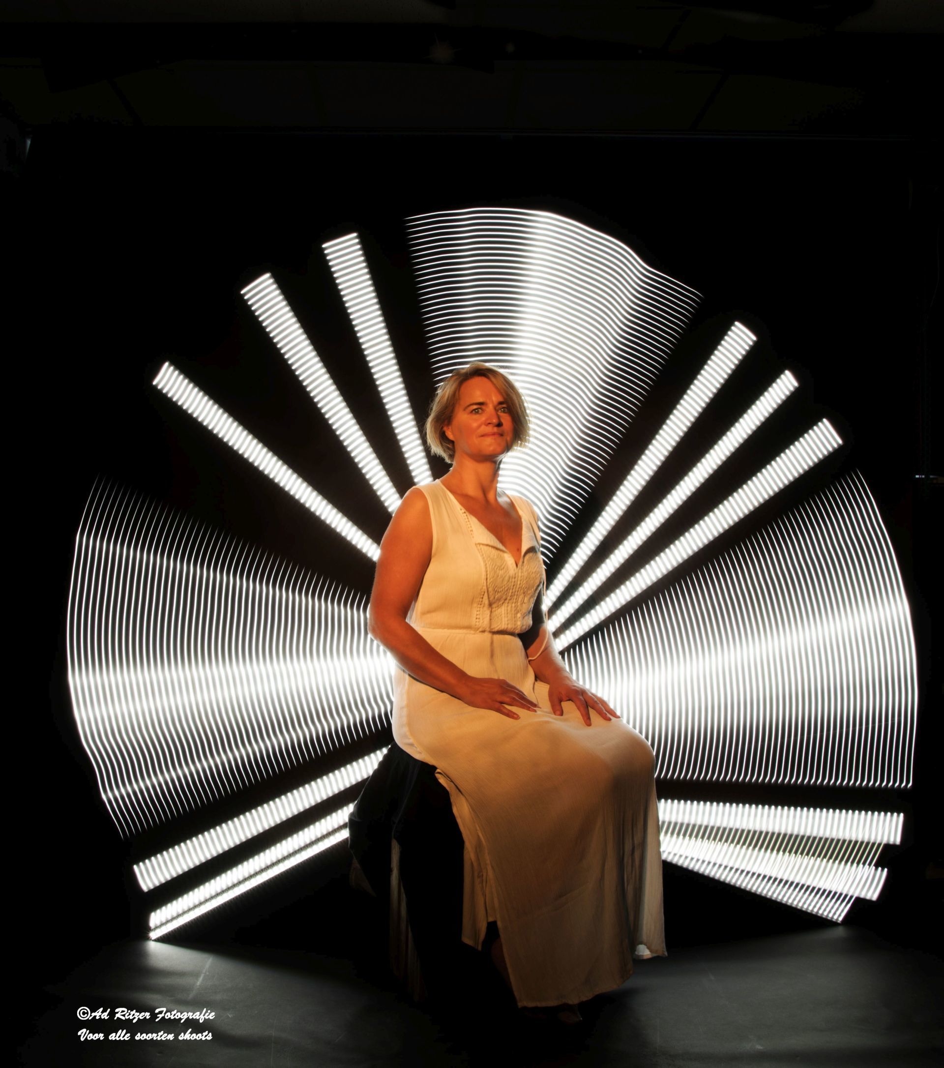 Vrouw in een witte jurk zit voor een zwart-witte achtergrond met radiale verlichting.