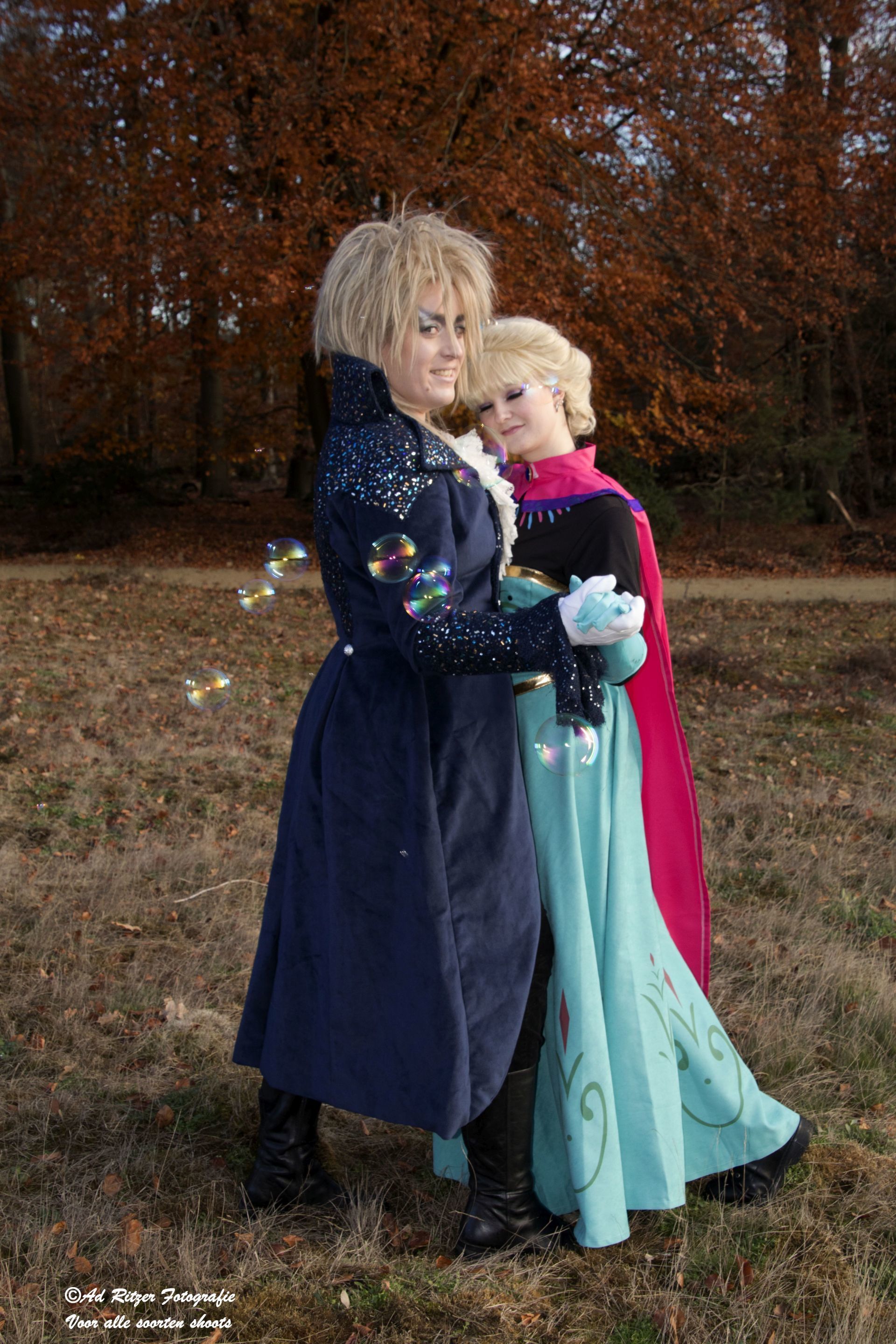 Twee personen in kostuum poseren buiten, gekleed in lange donkere en turquoise mantels, voor herfstbomen.