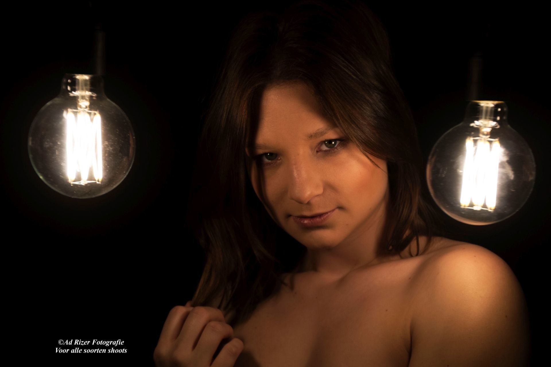 Vrouw, verlicht door twee hangende lampen tegen een donkere achtergrond, kijkt in de camera.