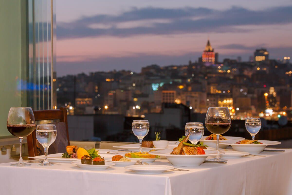A table with plates of food and wine glasses on it with a city in the background.