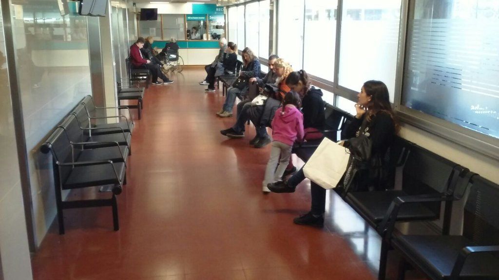 Personas esperando en bancos en un pasillo de hospital cerca de la recepción.