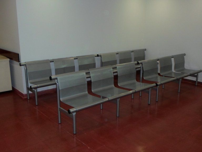 Bancos metálicos vacíos en una sala de espera con suelo rojo y pared blanca.