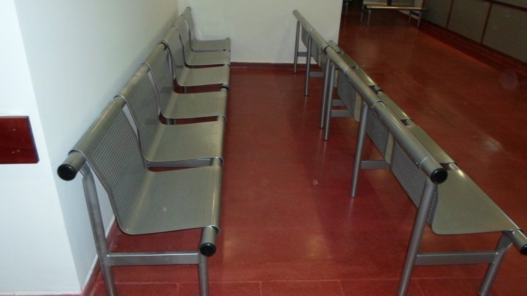 Sala de espera vacía con suelo rojo y bancos metálicos a lo largo de ambas paredes.
