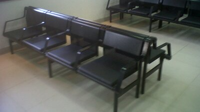 Banco de metal negro con tres asientos oscuros sobre un suelo de baldosas en el interior, con otro banco al fondo.