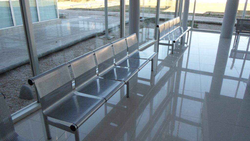 Una hilera de bancos metálicos de aeropuerto junto a grandes ventanales en un luminoso pasillo de la terminal.