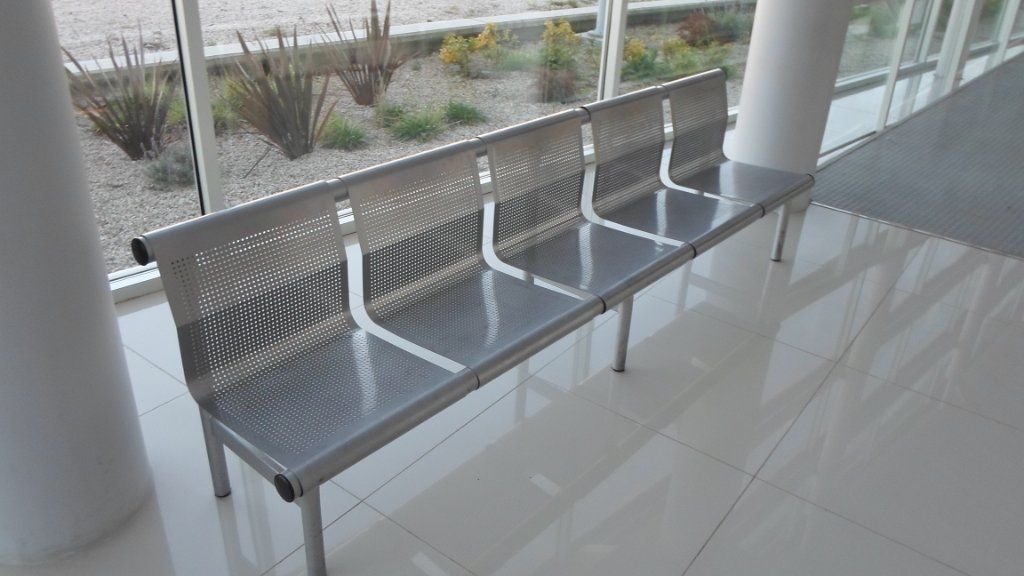 Hilera de bancos de metal plateado en una pasarela cubierta de baldosas brillantes con vistas a una zona ajardinada.